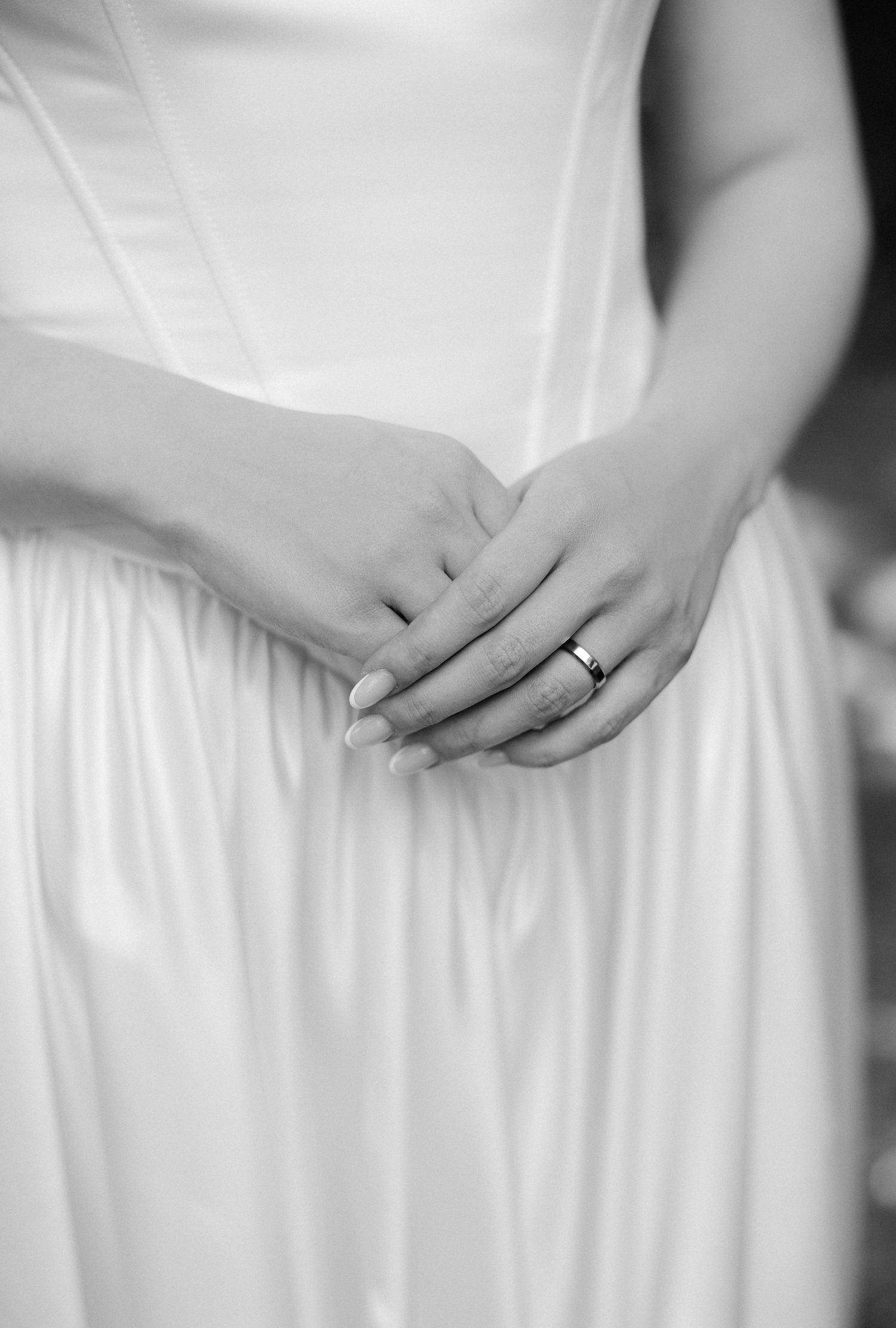 Close-up black and white photo of bride's hands clasped together wearing a wedding band on white gown.