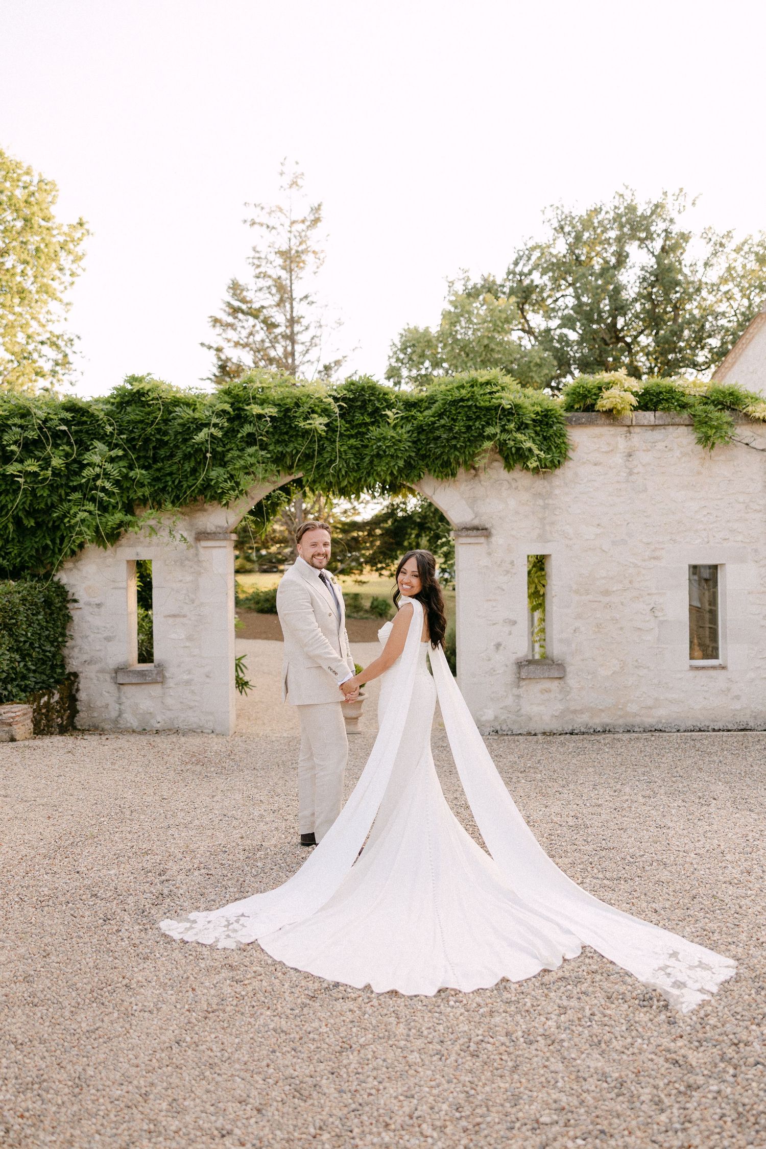 Bride and groom holding hands before ivy-covered stone archway at golden hour wedding portrait.