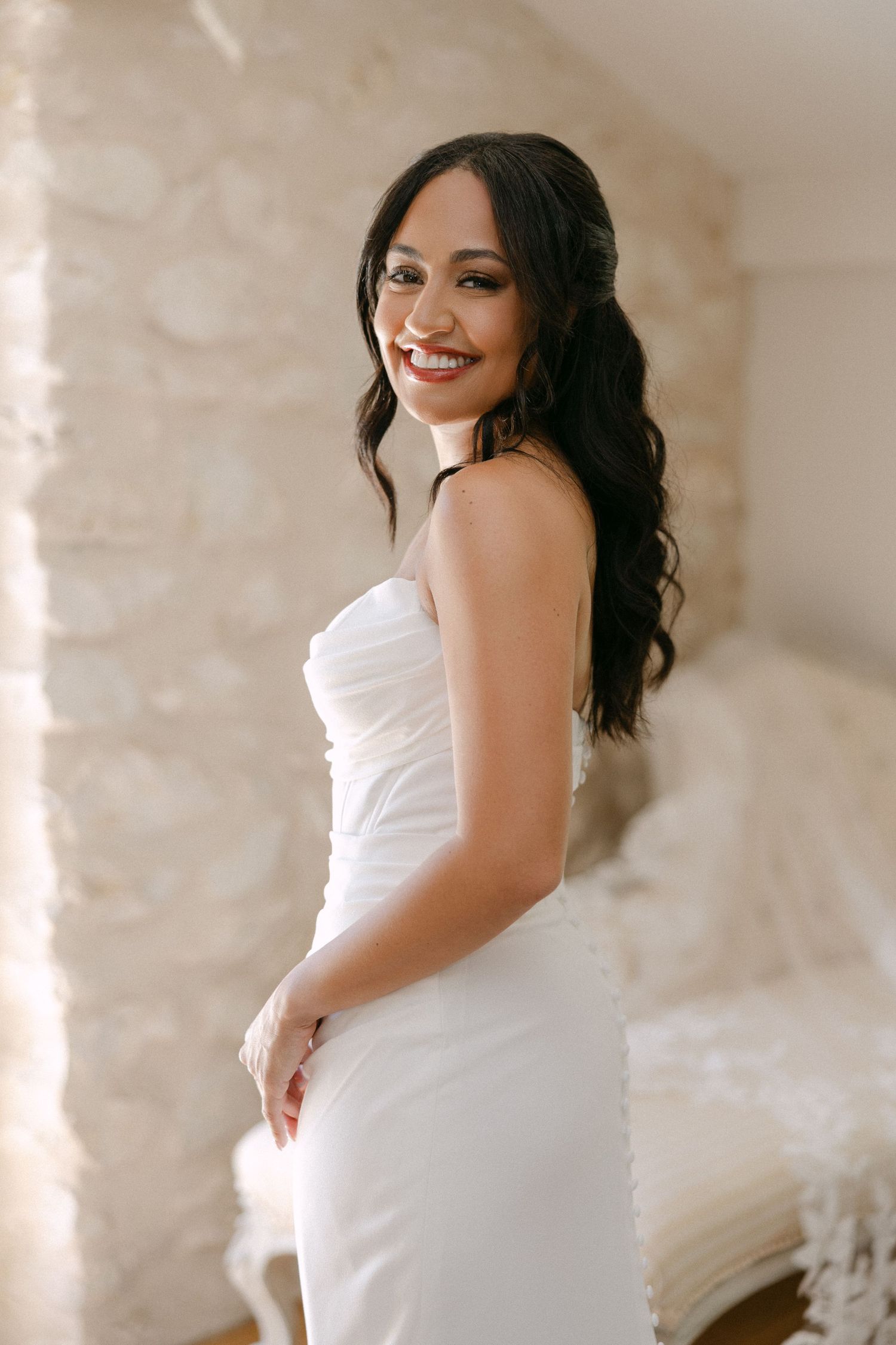 Smiling dark-haired bride in strapless white wedding dress posing in bright stone-walled bridal suite.
