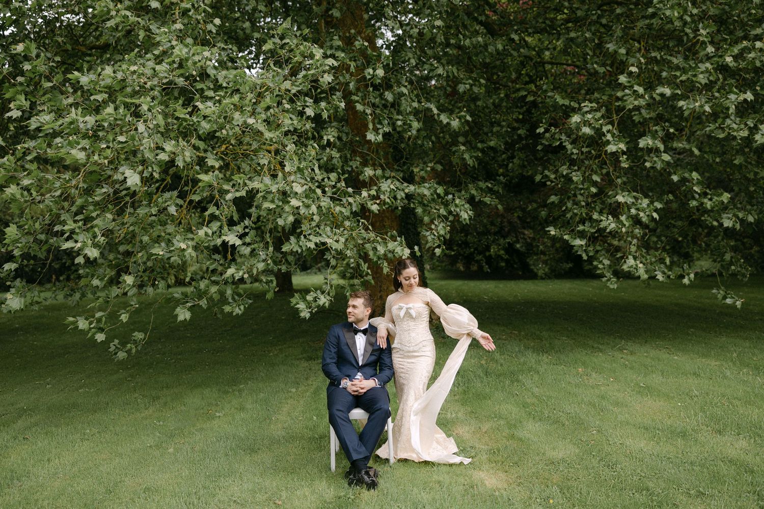 Bride and groom posing on green lawn beneath large tree at elegant outdoor wedding venue.