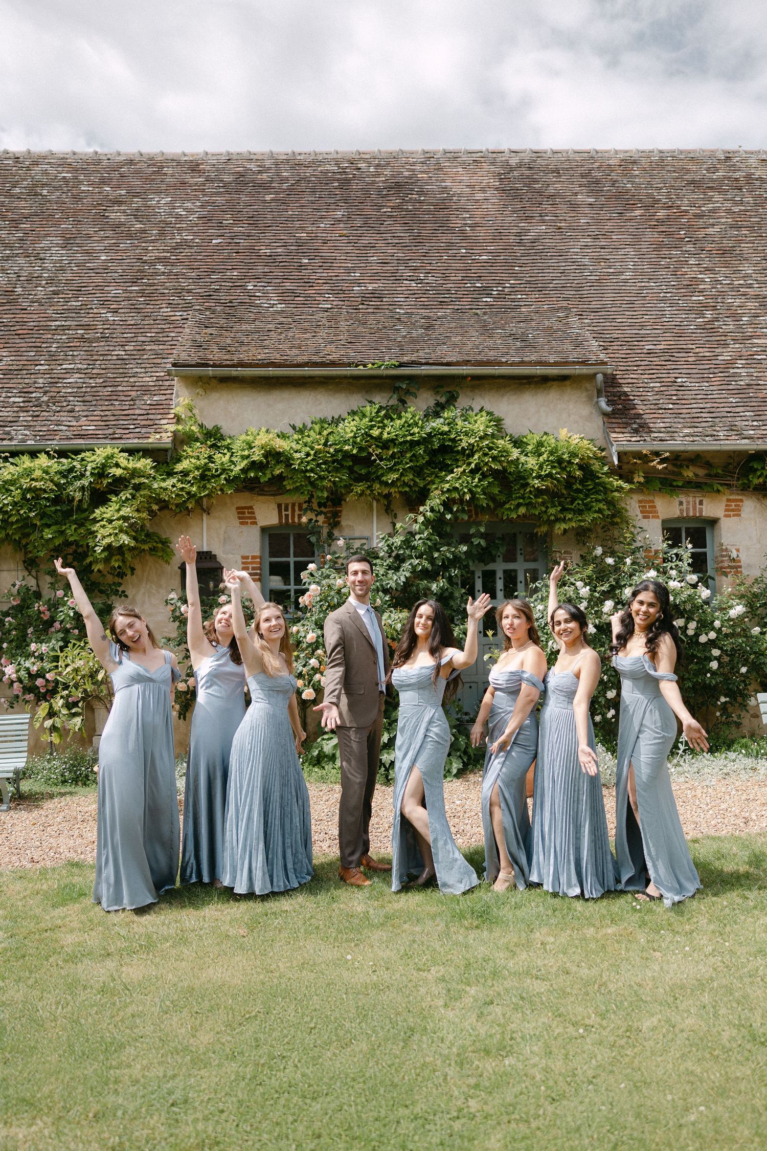 Groom in brown suit poses with six bridesmaids in dusty blue gowns outside a charming French estate.