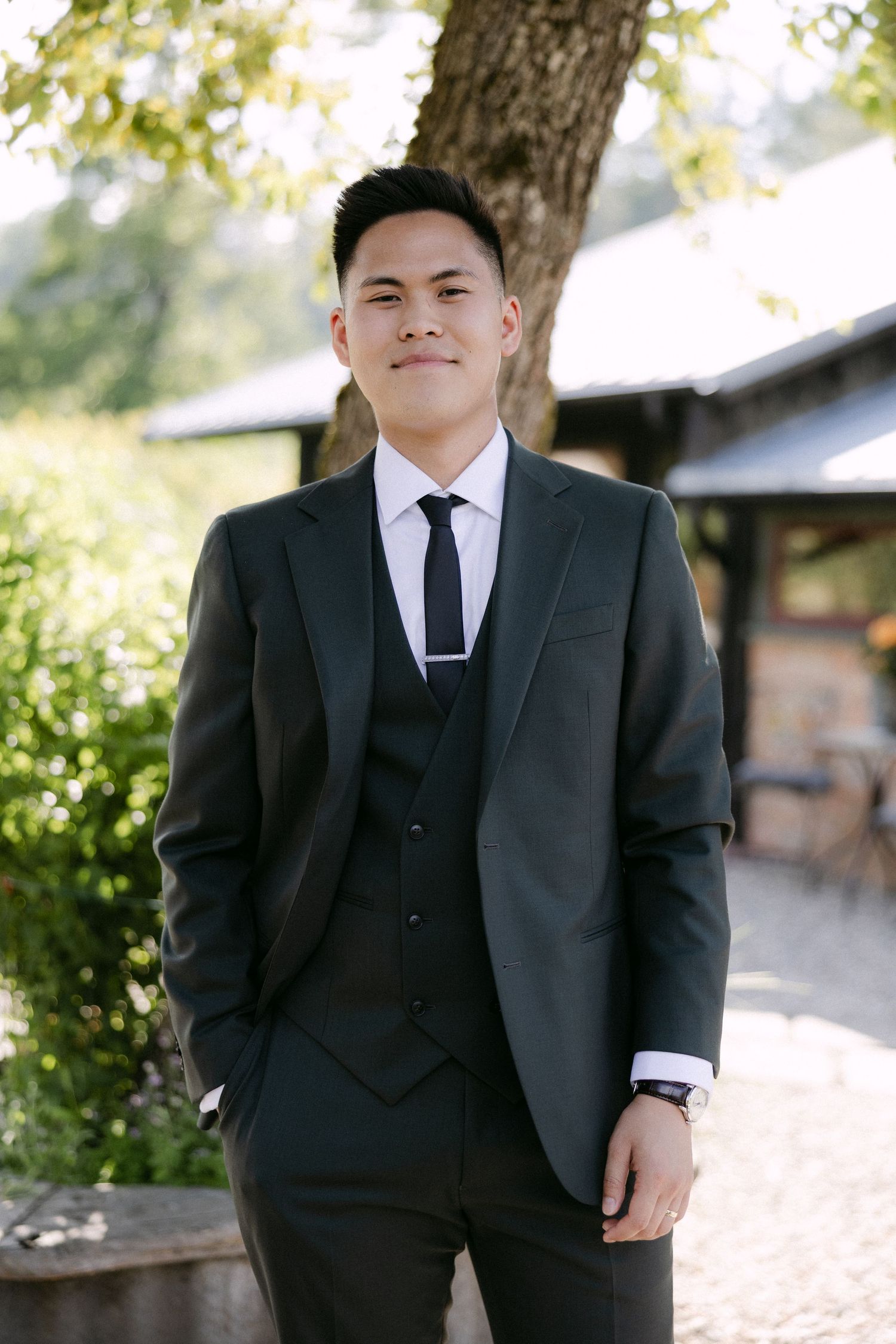 Young Asian man in dark three-piece suit with black tie poses outdoors at wedding venue.