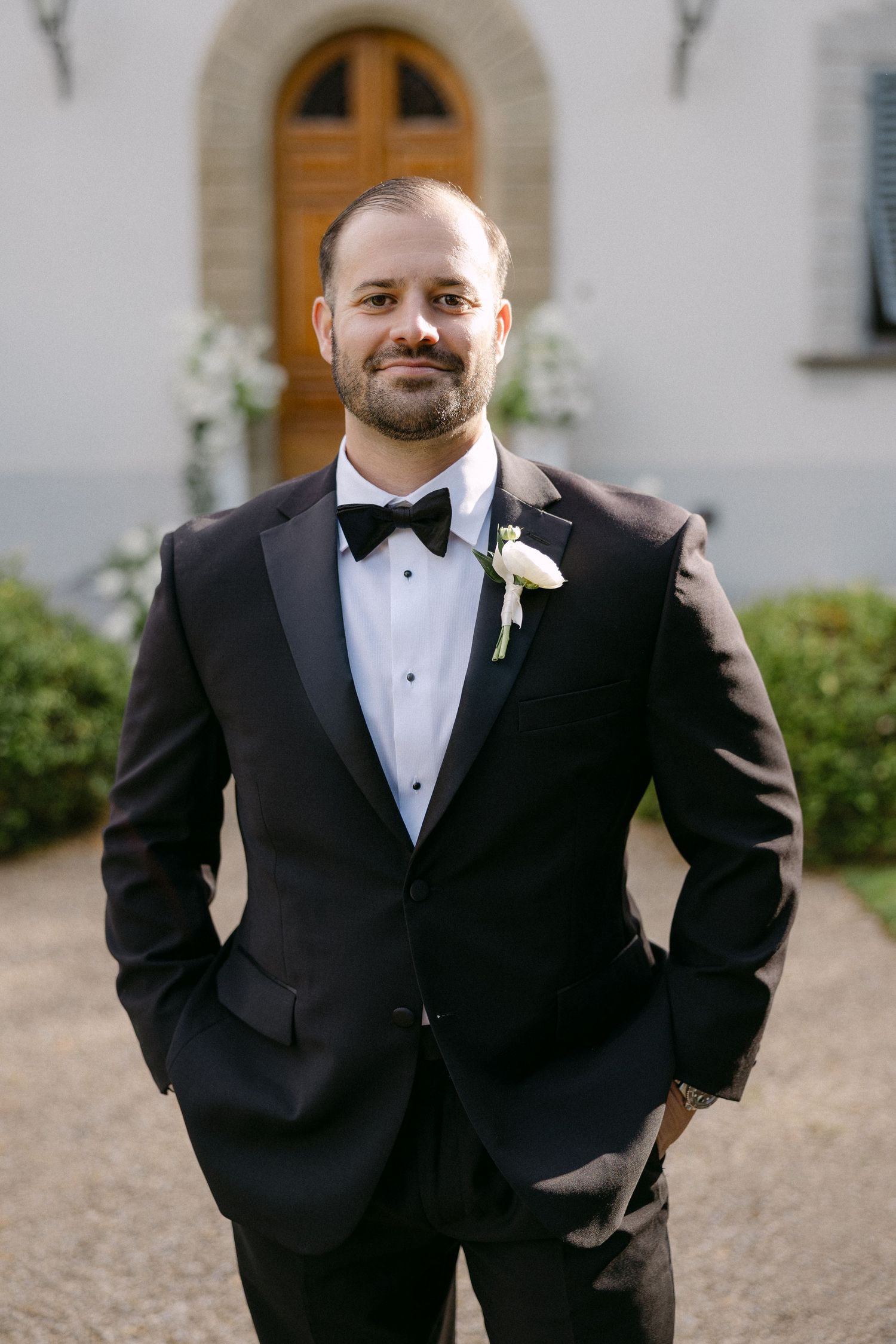 Dapper groom in black tuxedo with bow tie and white boutonniere stands confidently outside a church entrance.