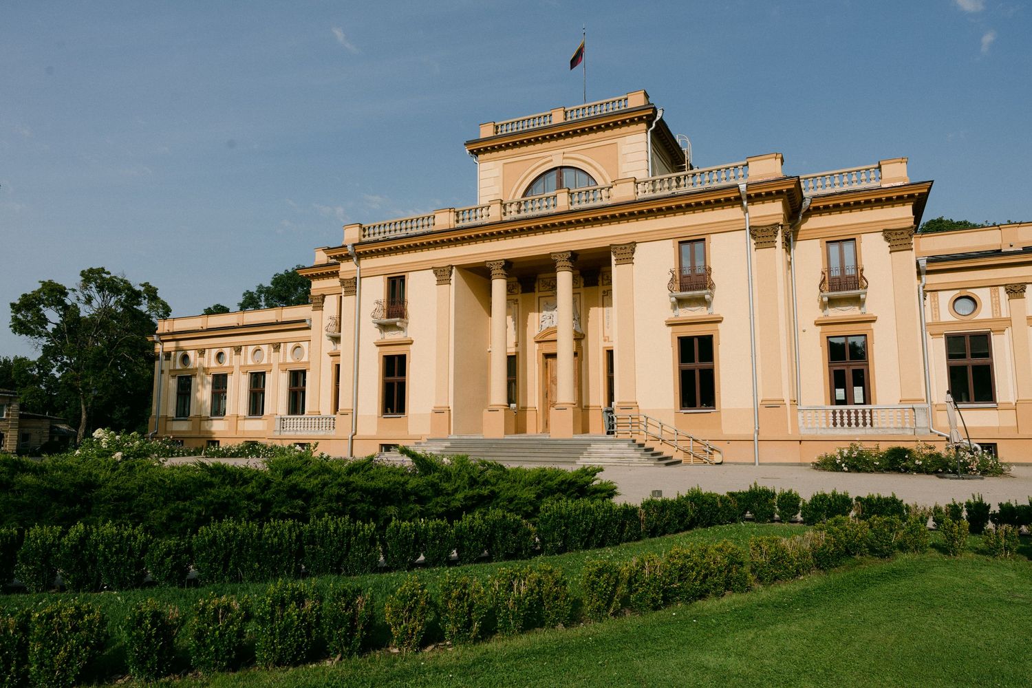 Grand neoclassical palace with columns, manicured hedges, and a flag on a clear sunny day.