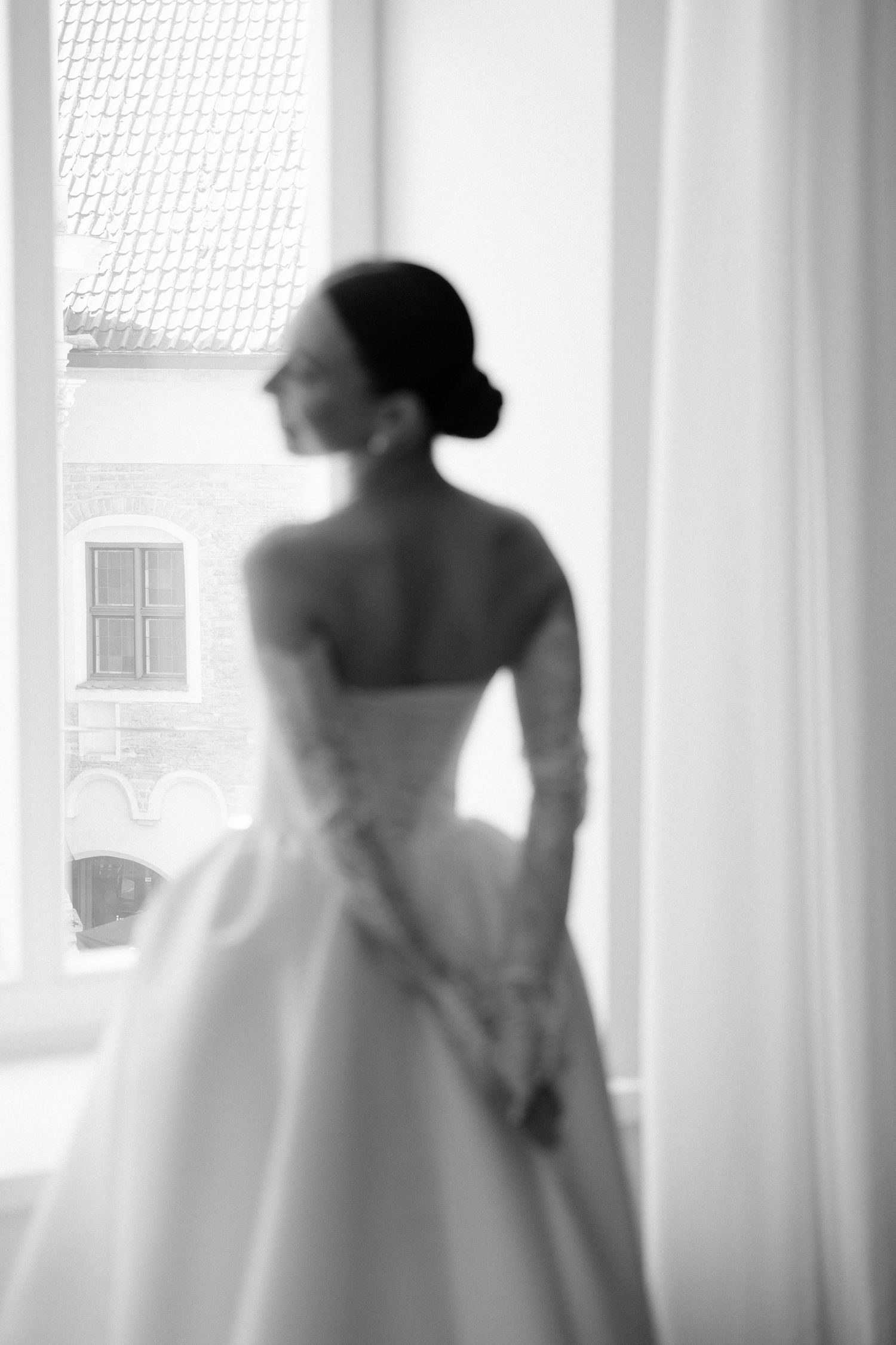 Elegant bride in strapless white ball gown with lace gloves stands by window in soft black and white photo.