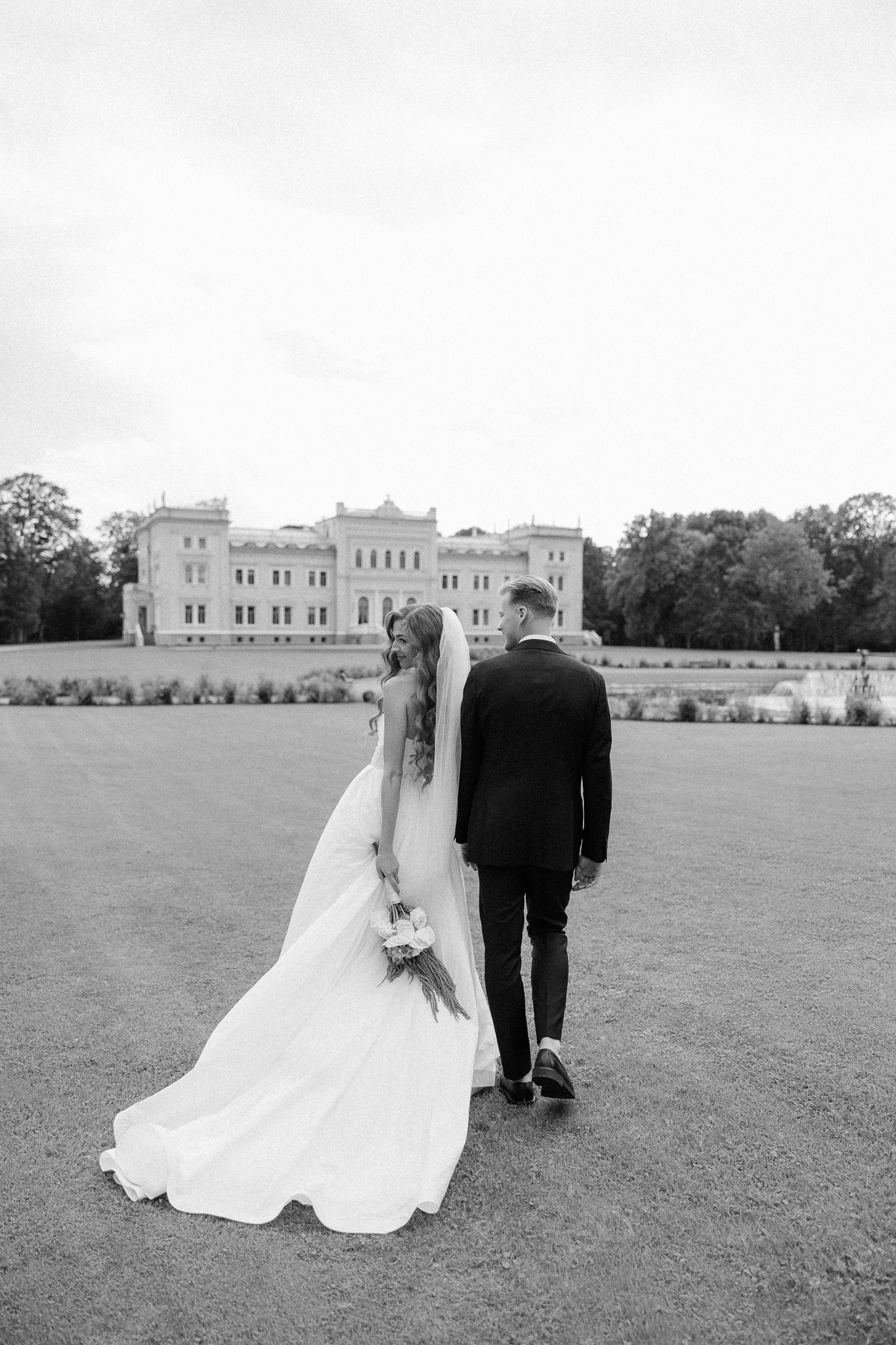 A couple in wedding attire stands together on a vast lawn in front of a grand stone castle-like building.