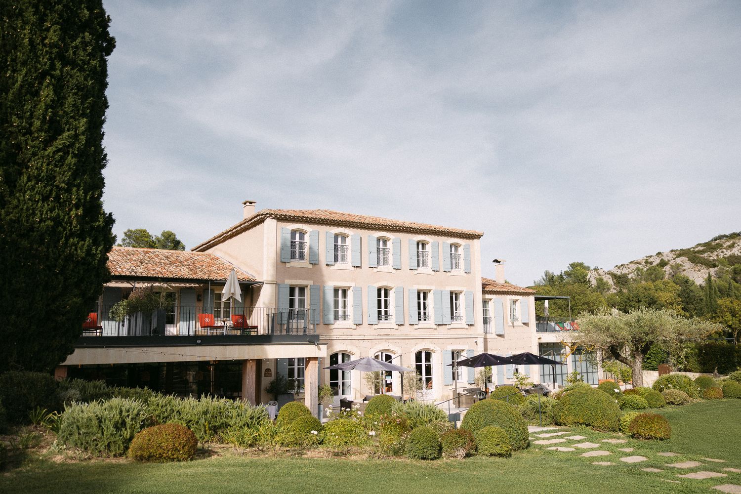Elegant Provençal villa with manicured gardens, cypress trees, and mountain backdrop under a clear blue sky.