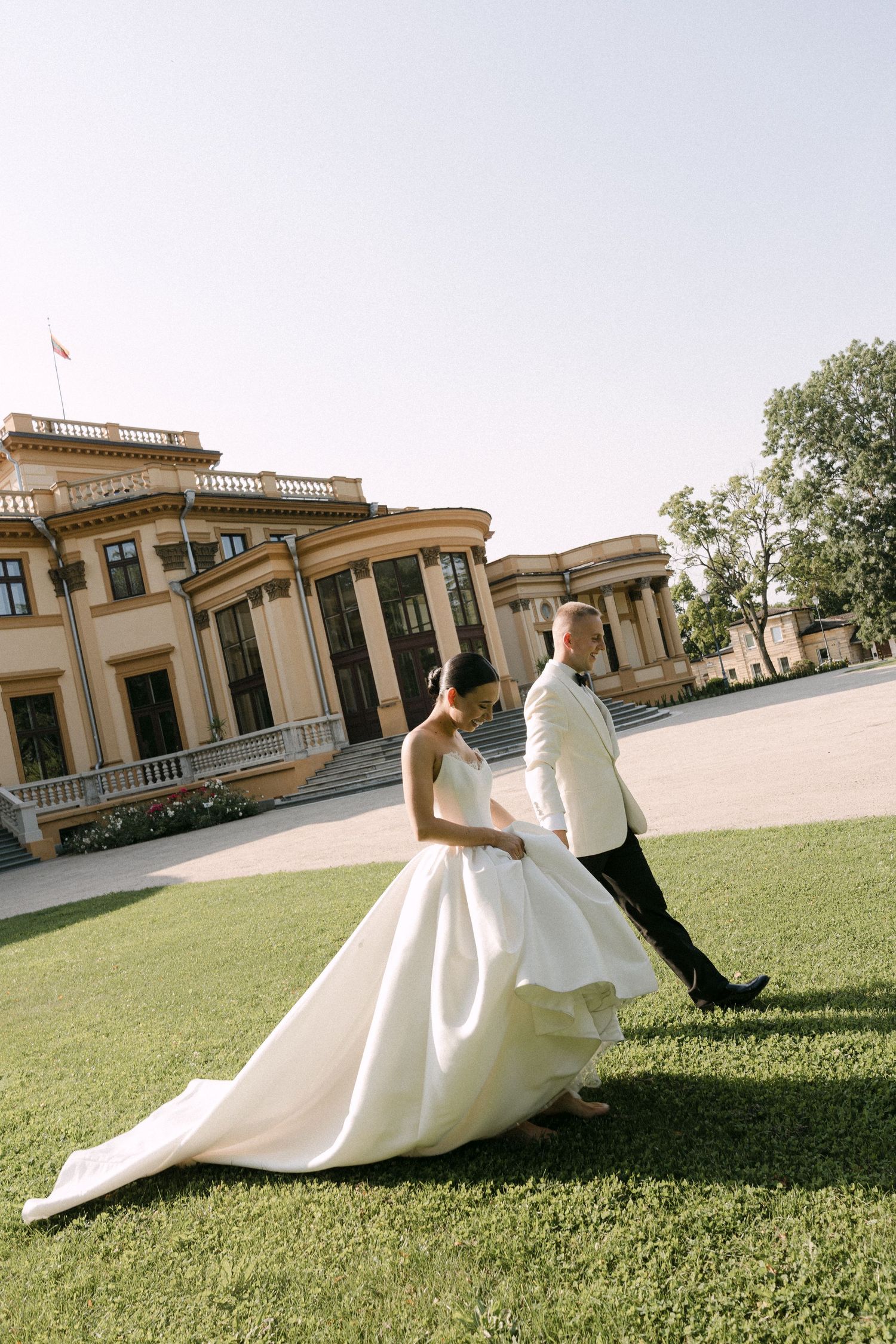 Bride in flowing white gown and groom in white tuxedo walking on green lawn before grand estate.