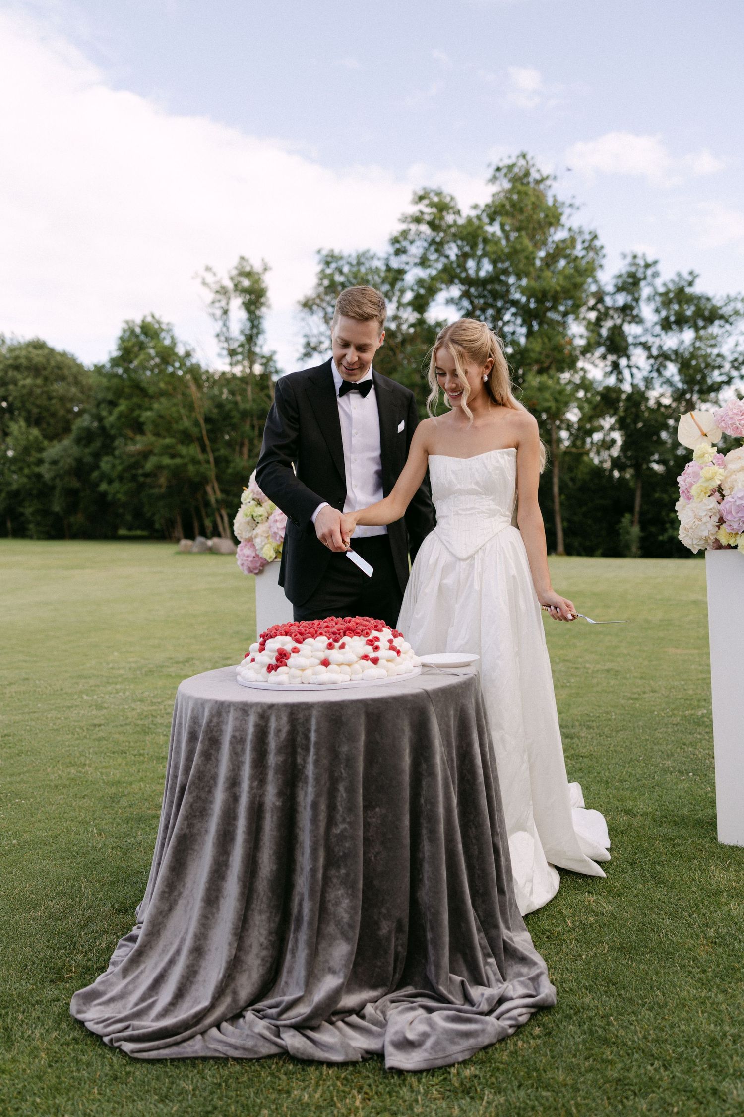 Bride and groom cut their elegant white wedding cake topped with red berries on an outdoor lawn.