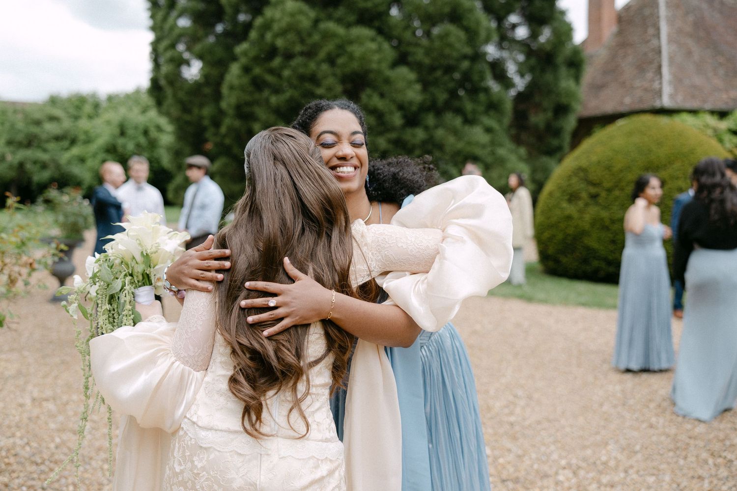 Bride in white dress joyfully embraces a smiling guest at outdoor wedding celebration.
