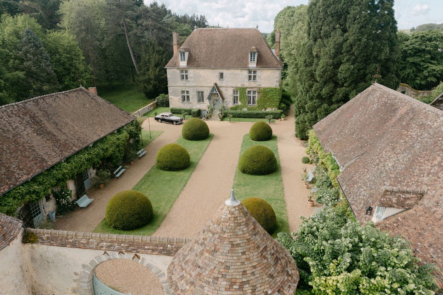 Aerial view of a French manor house with manicured topiary gardens and terracotta-roofed outbuildings.