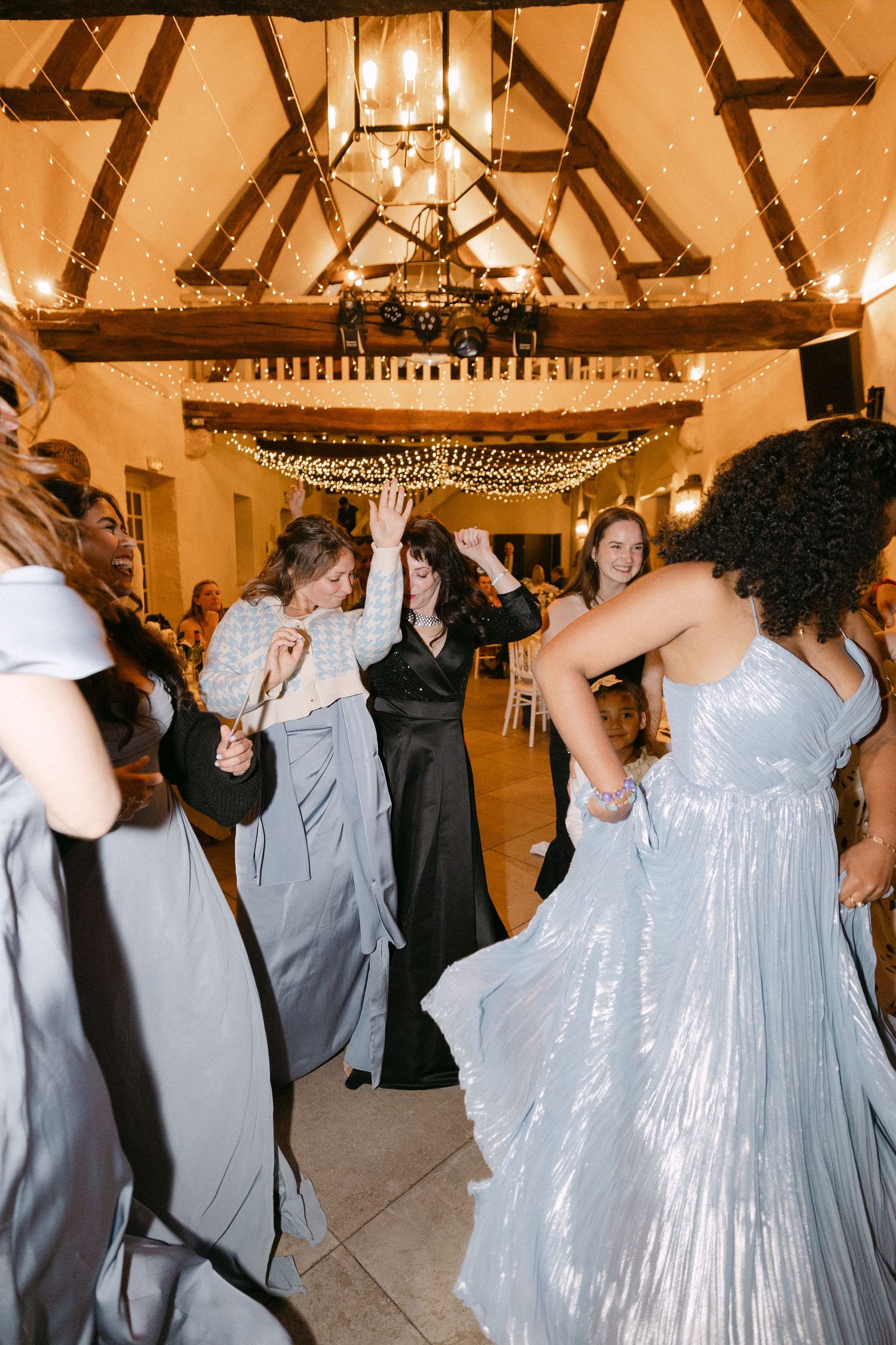 Elegant wedding guests in silver gowns dancing joyfully under fairy lights in a decorated barn venue.