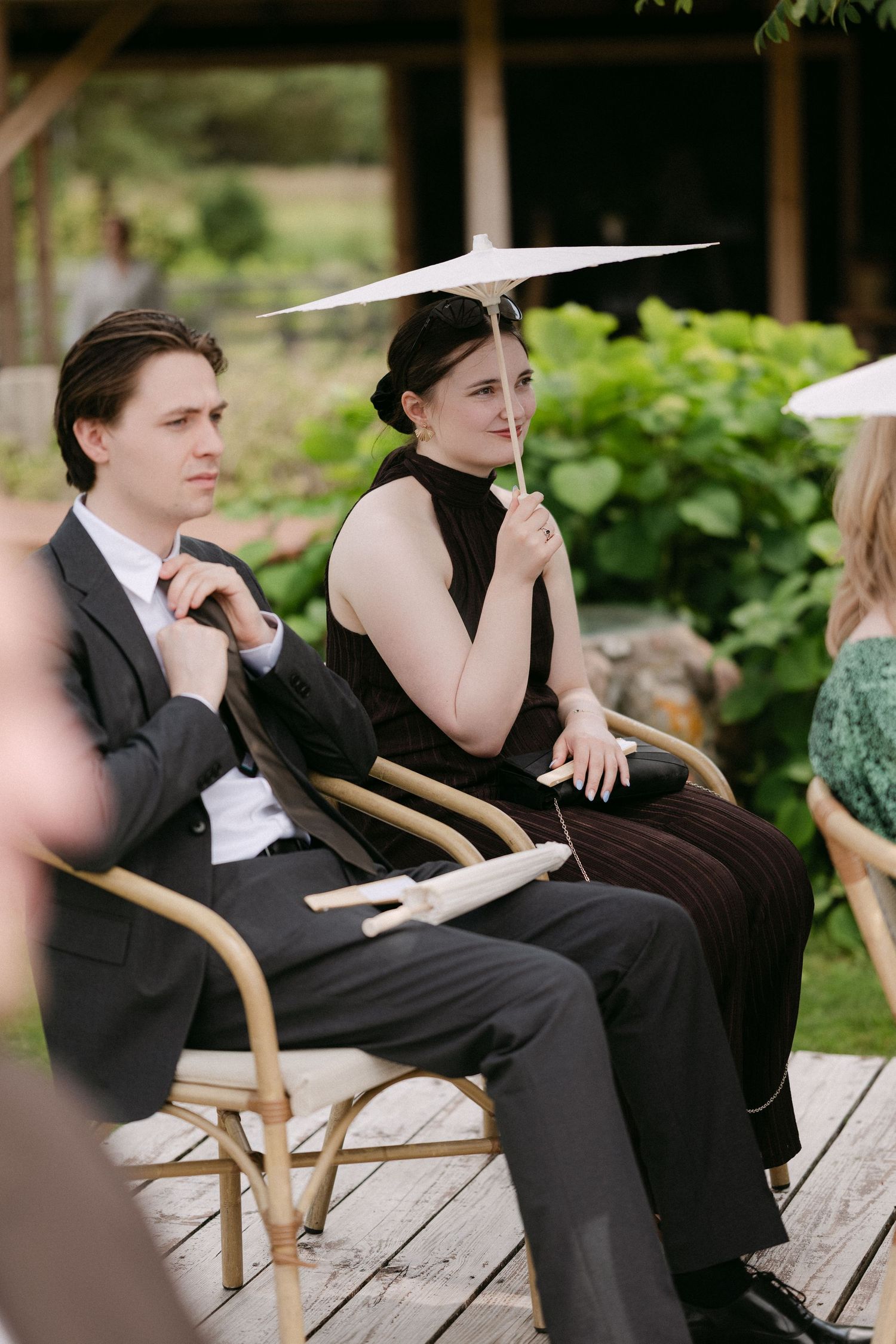 Wedding guests sit in rattan chairs outdoors, woman holding white parasol umbrella at garden ceremony.