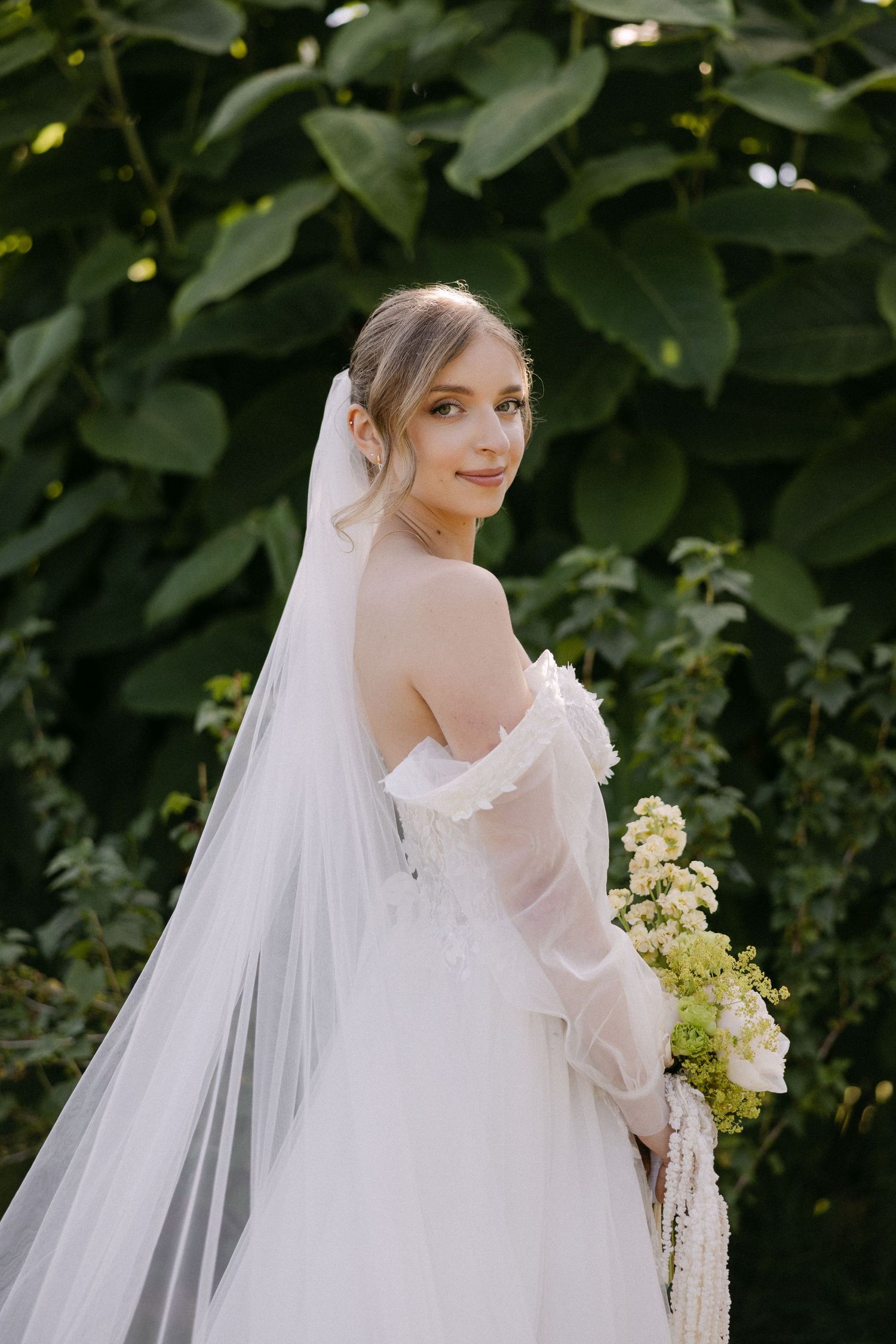 Elegant bride in off-shoulder lace wedding gown with long veil holding bouquet against green foliage.
