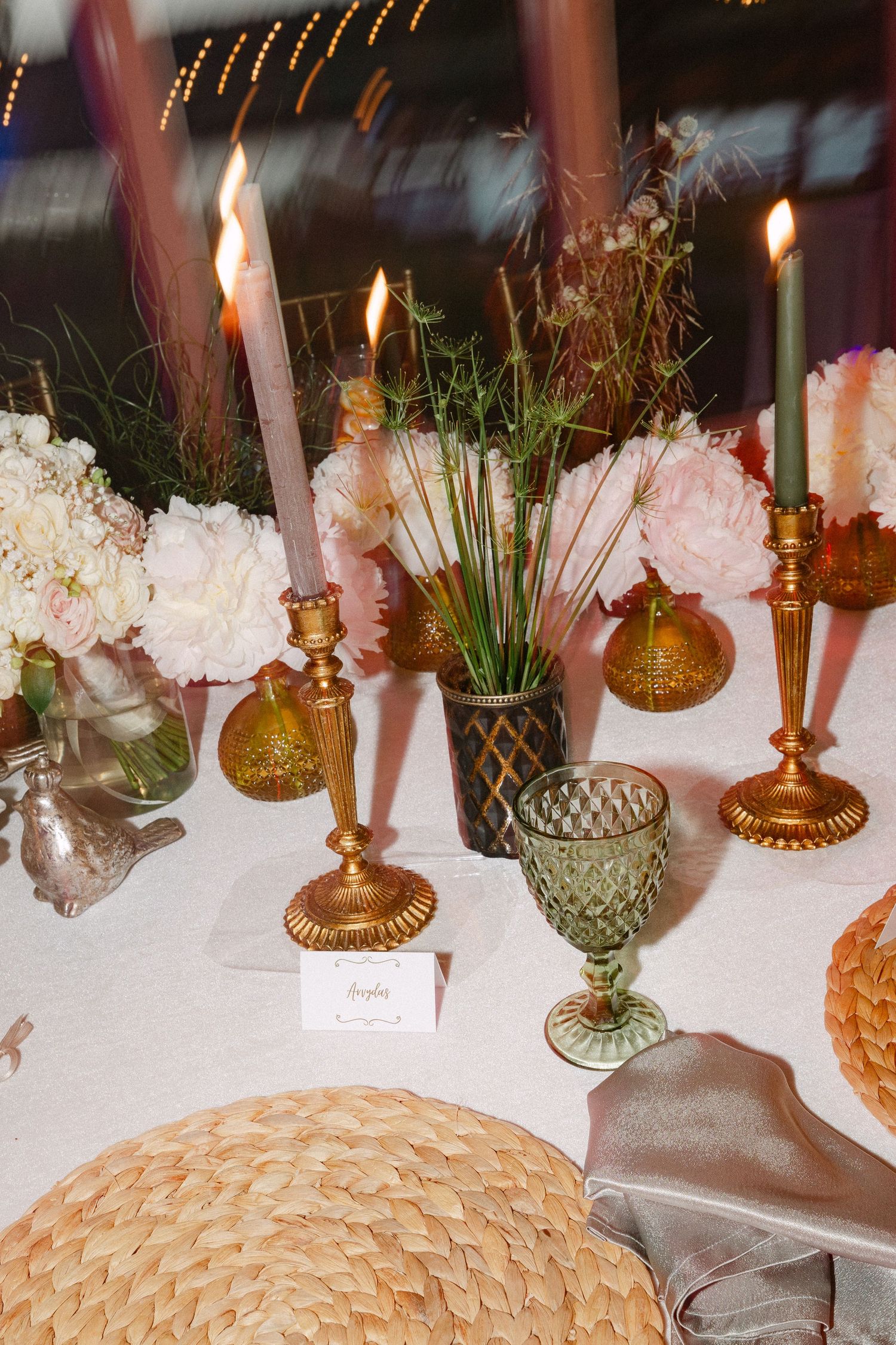 Elegant wedding tablescape with gold candlesticks, pink peonies, green glassware, and warm candlelight.