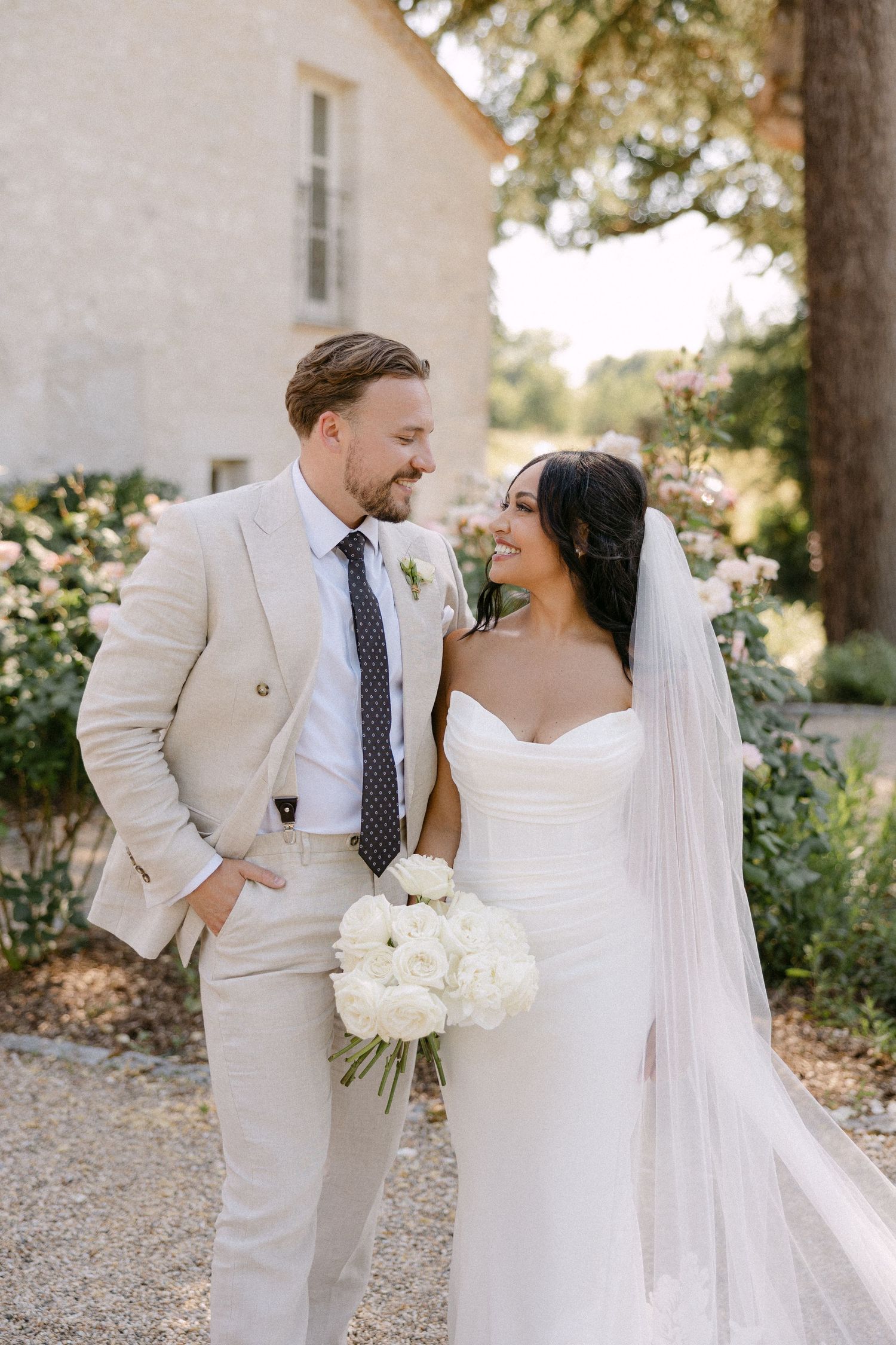 Bride and groom smiling at each other outdoors, she holds white bouquet in wedding dress with veil, he wears beige suit.
