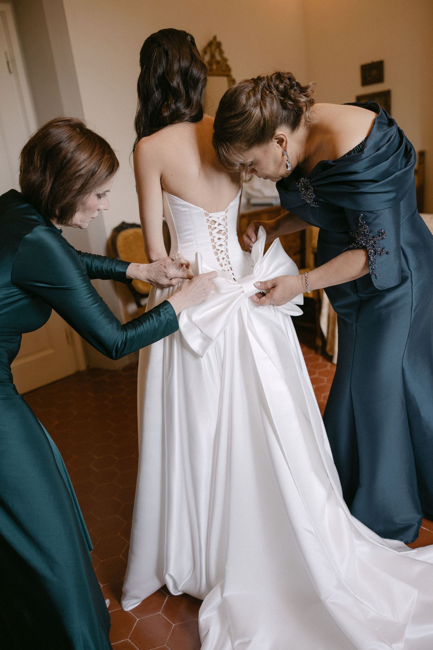 Two bridesmaids in green and blue gowns help bride lace up her white strapless wedding dress with bow detail.