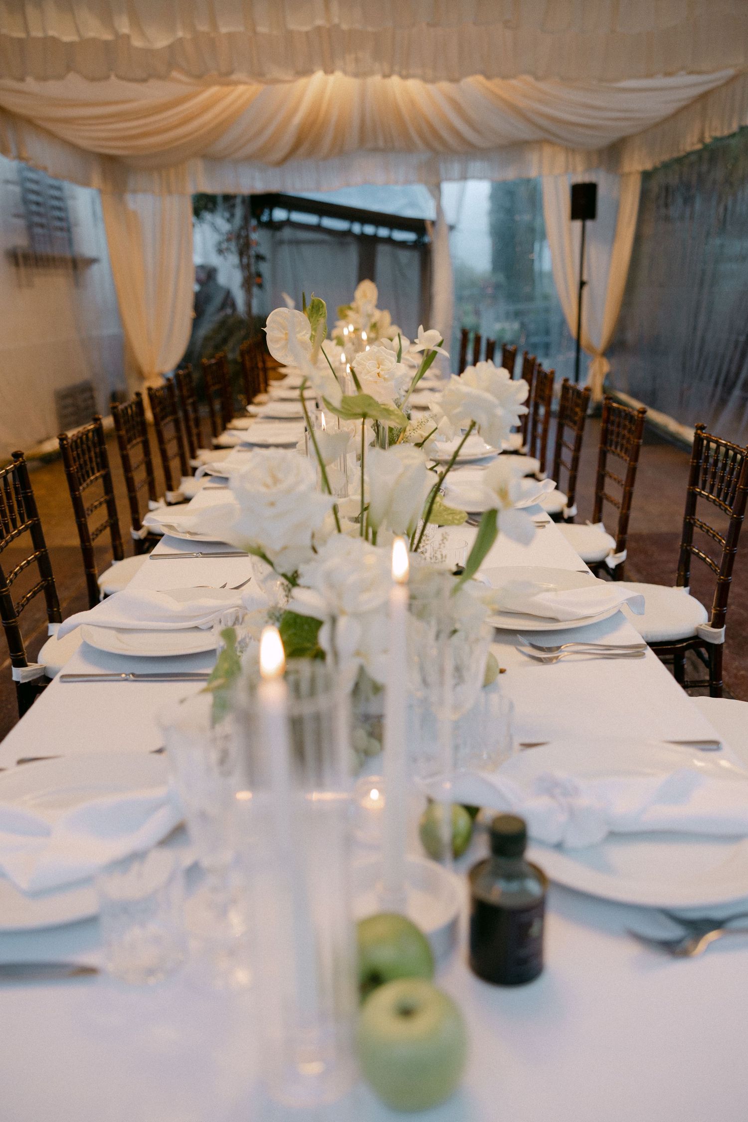 Elegant wedding reception table set with white florals, candles, and green apples under draped tent.