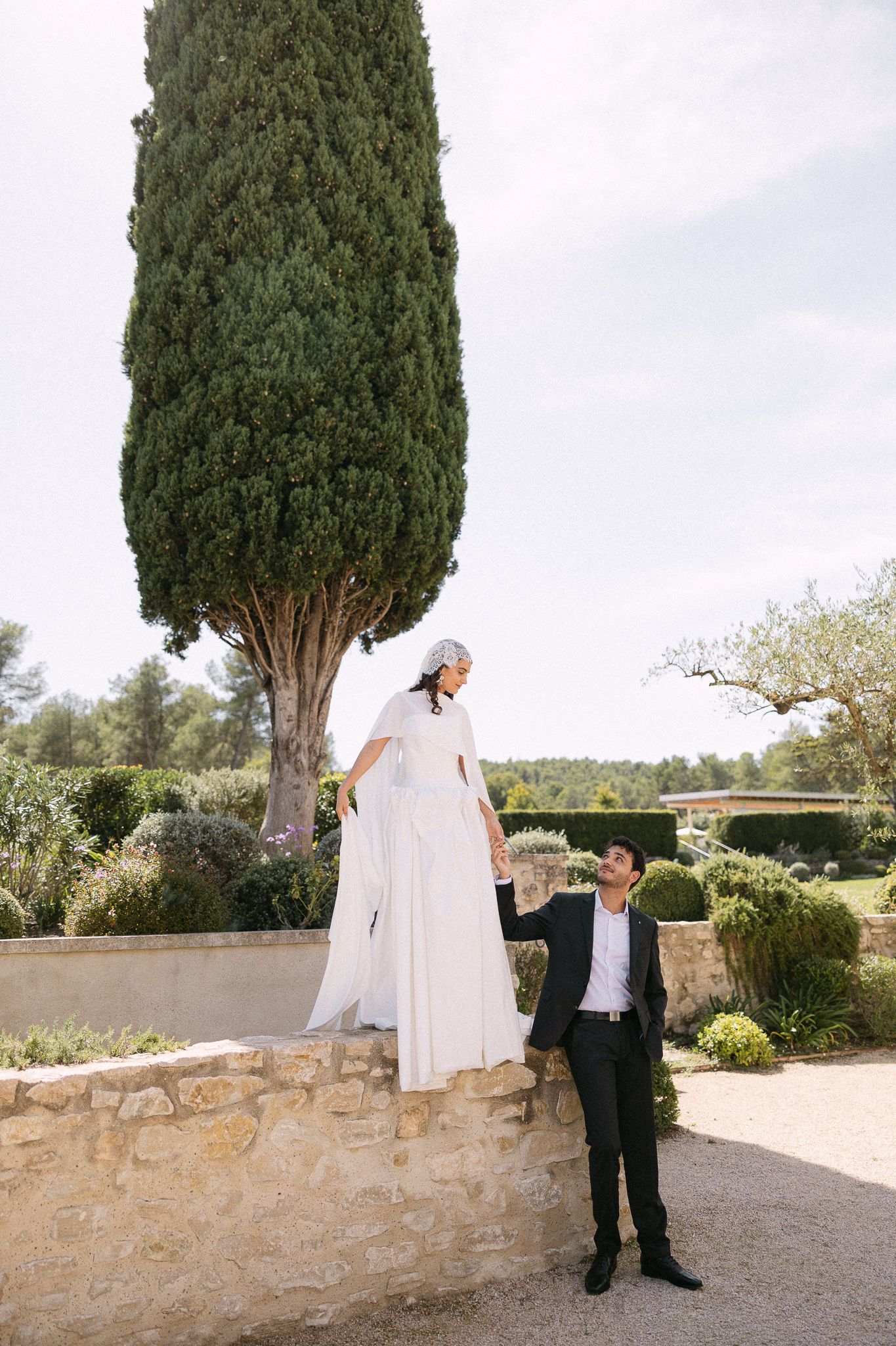 Bride in white gown stands on stone wall holding groom's hand in lush garden with tall cypress tree.