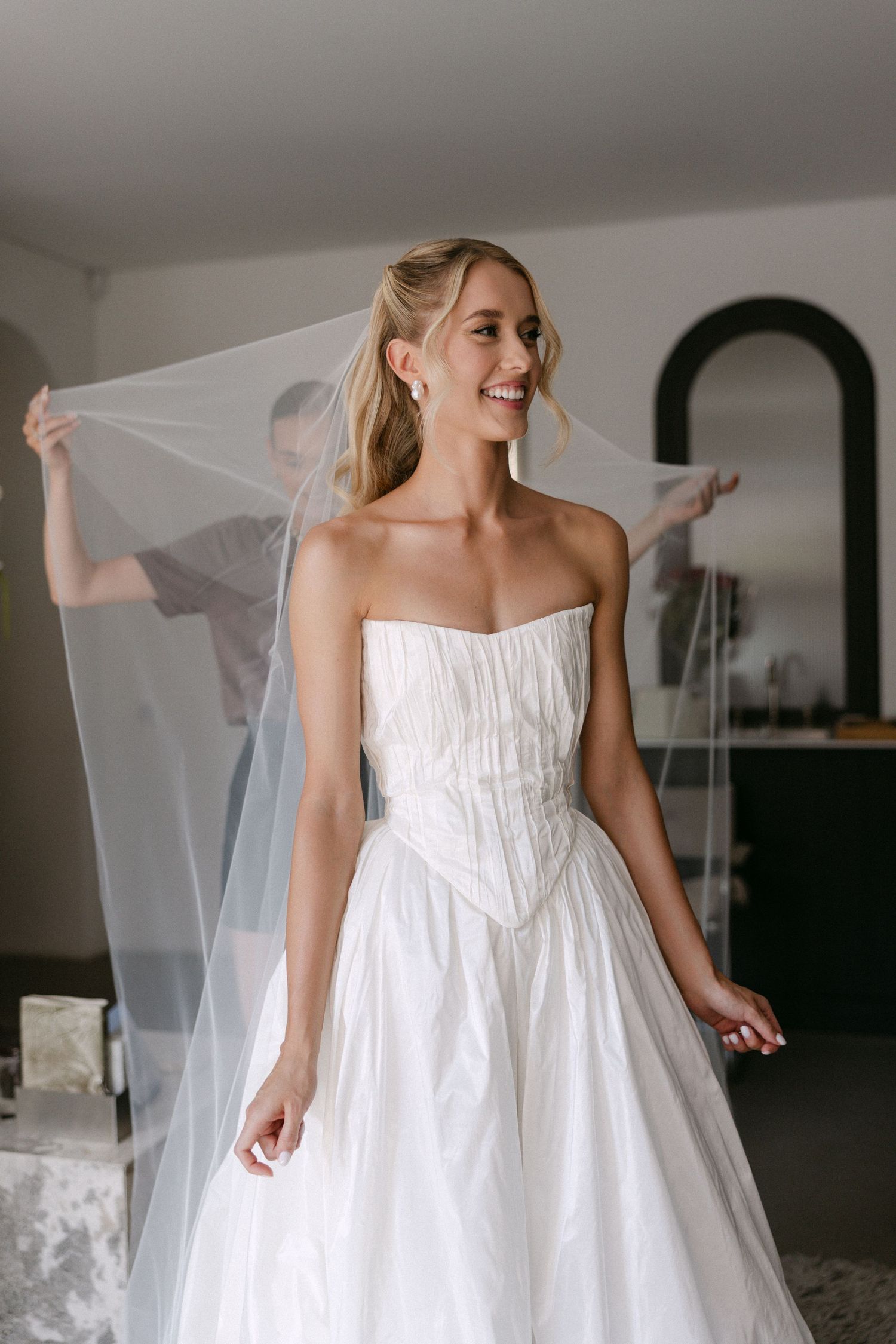 Smiling blonde bride in strapless white ball gown with veil being arranged by assistant indoors.