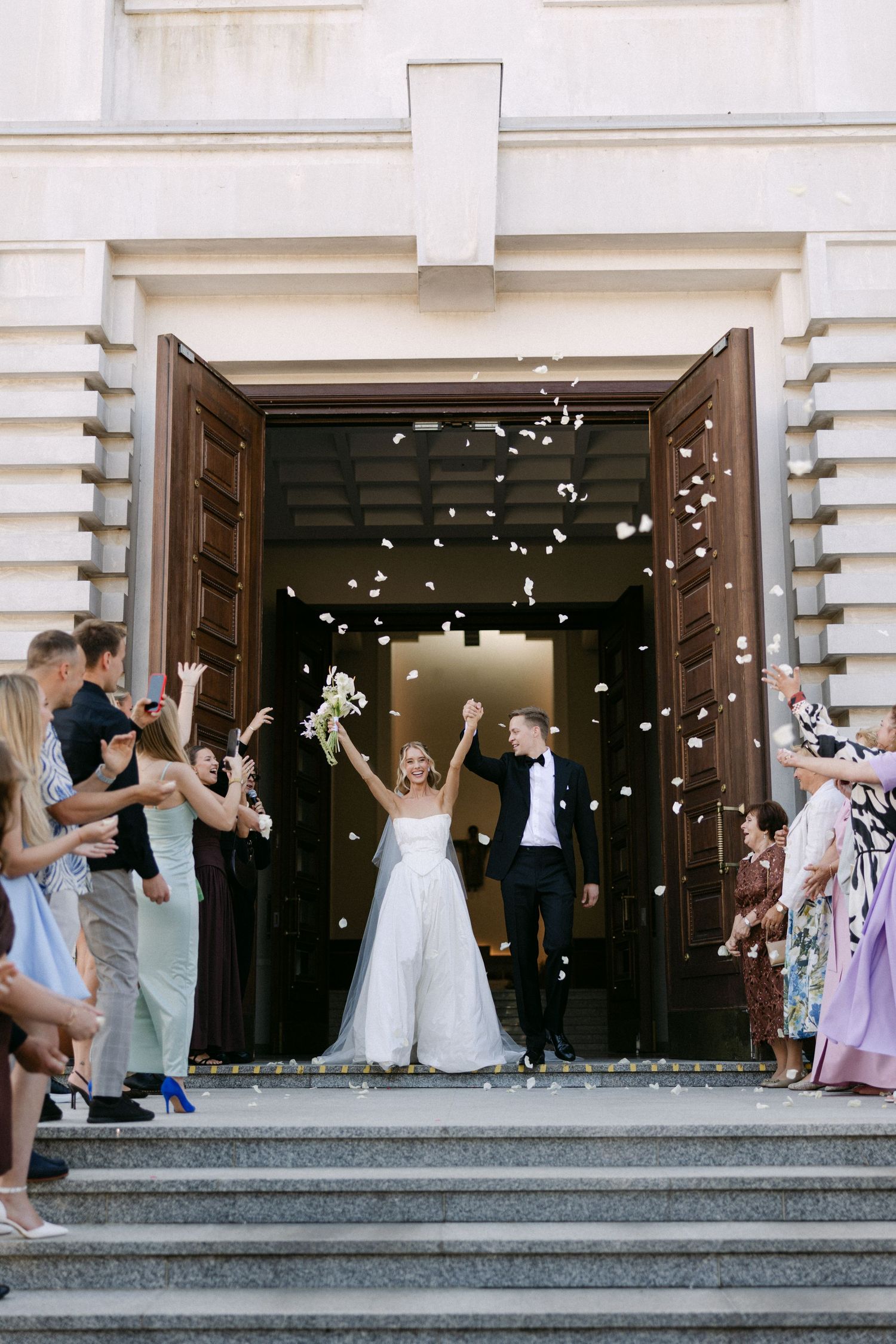 Bride and groom exit church doors as guests toss white confetti petals in joyful wedding celebration.