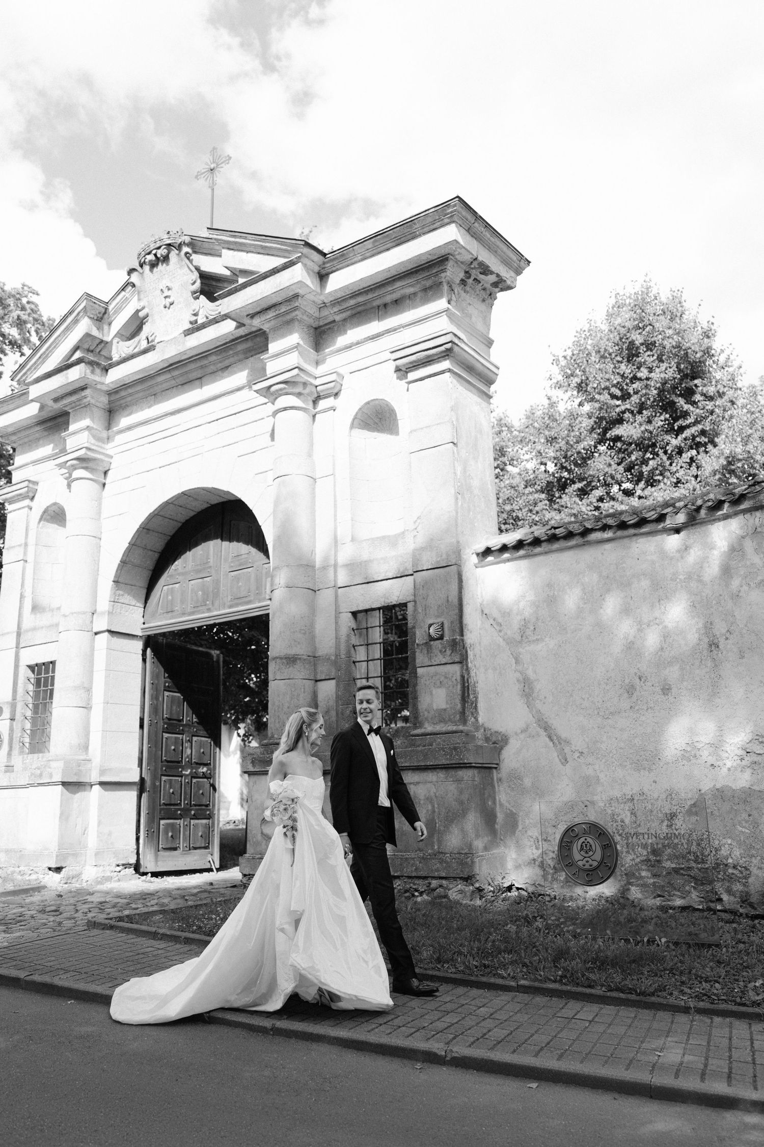 Bride and groom walking hand in hand before an ornate stone archway in black and white photo.