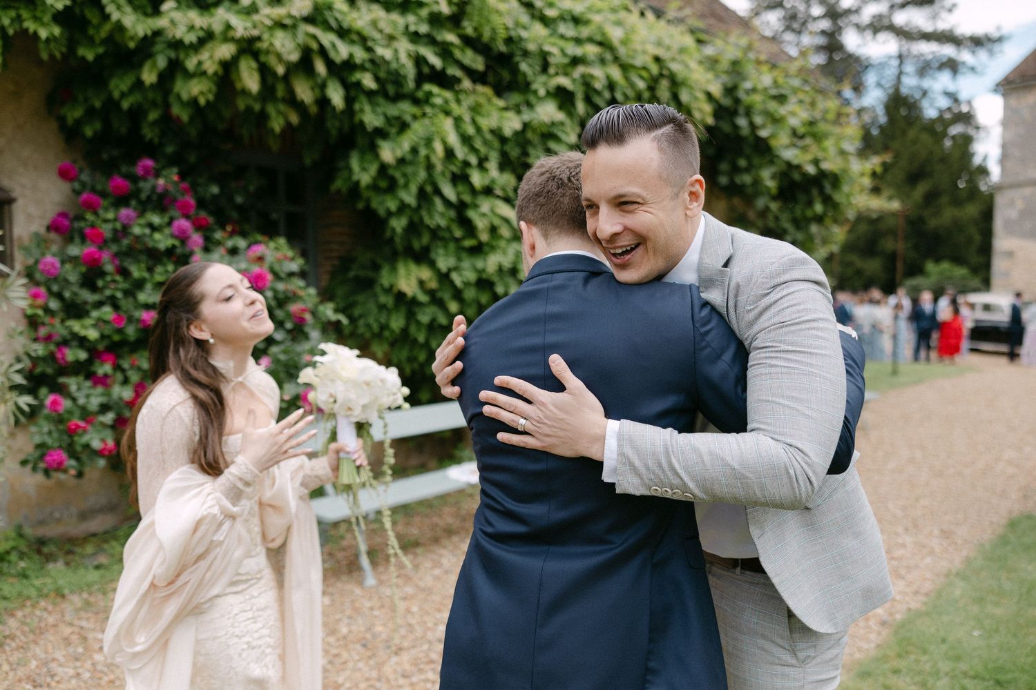 Groom in navy suit embracing a guest while bride in white dress holds bouquet at wedding venue.