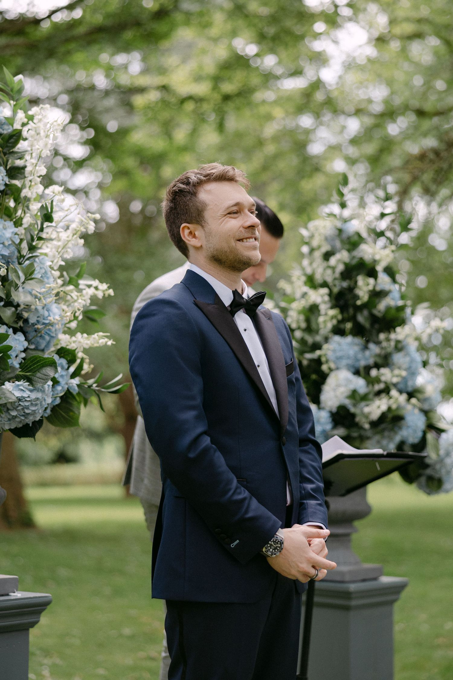 Smiling groom in navy tuxedo standing at outdoor wedding altar surrounded by blue hydrangea floral arrangements.