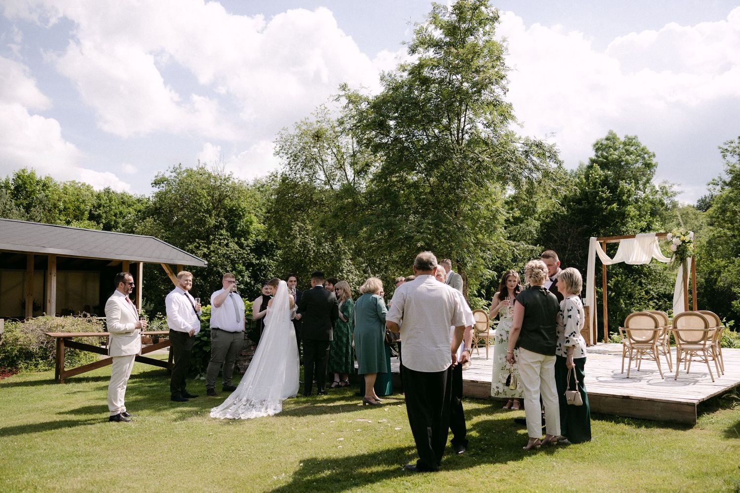 Wedding guests mingle on sunny lawn with bride in white gown near rustic outdoor ceremony space.