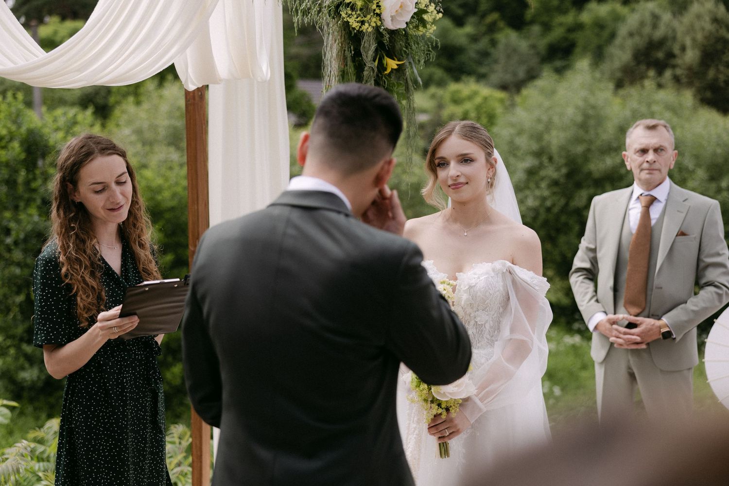 Bride smiles at groom during outdoor wedding ceremony with officiant and guest beside floral arch.