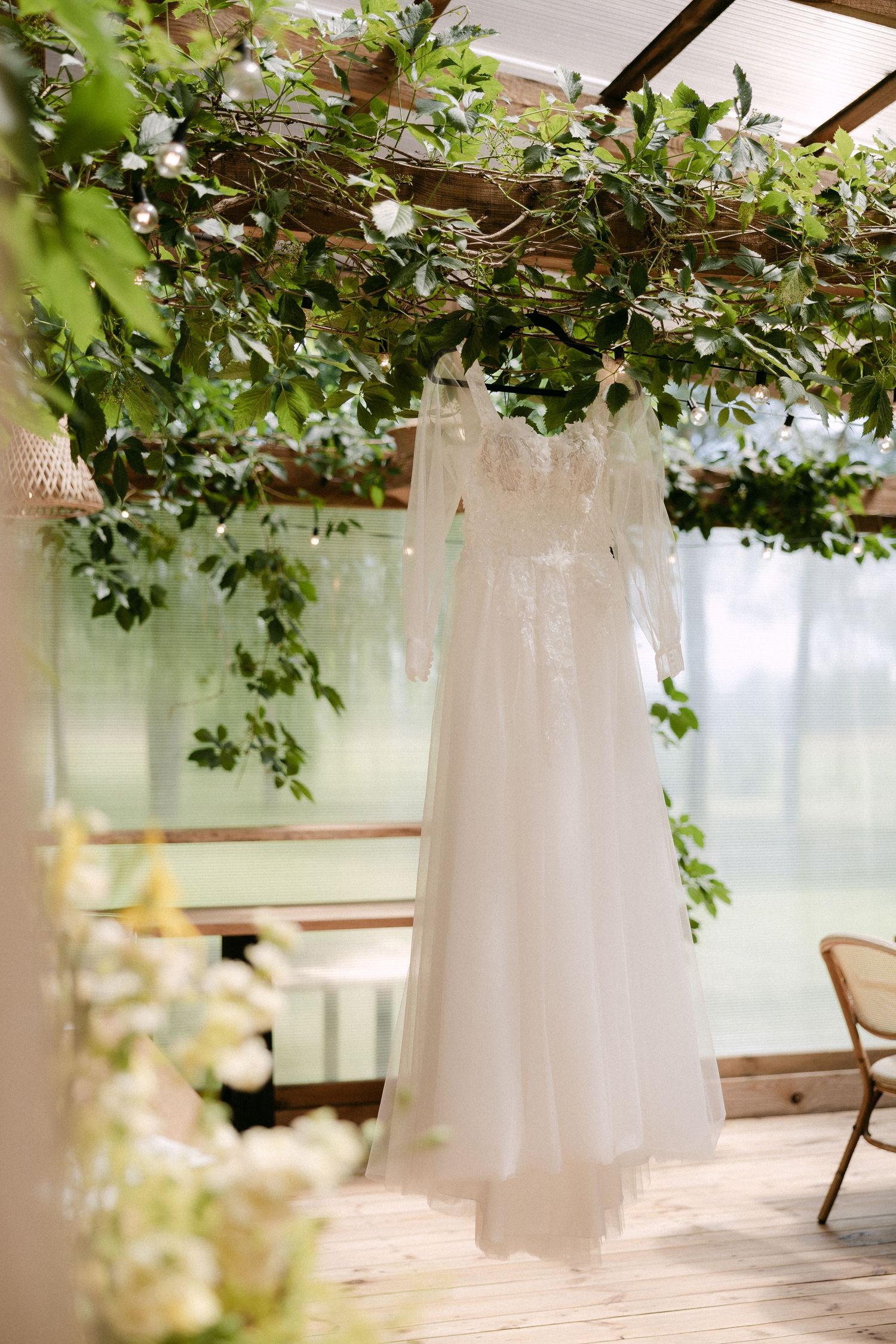 Elegant lace wedding dress with long sleeves hanging among green floral decorations in a sunlit greenhouse.
