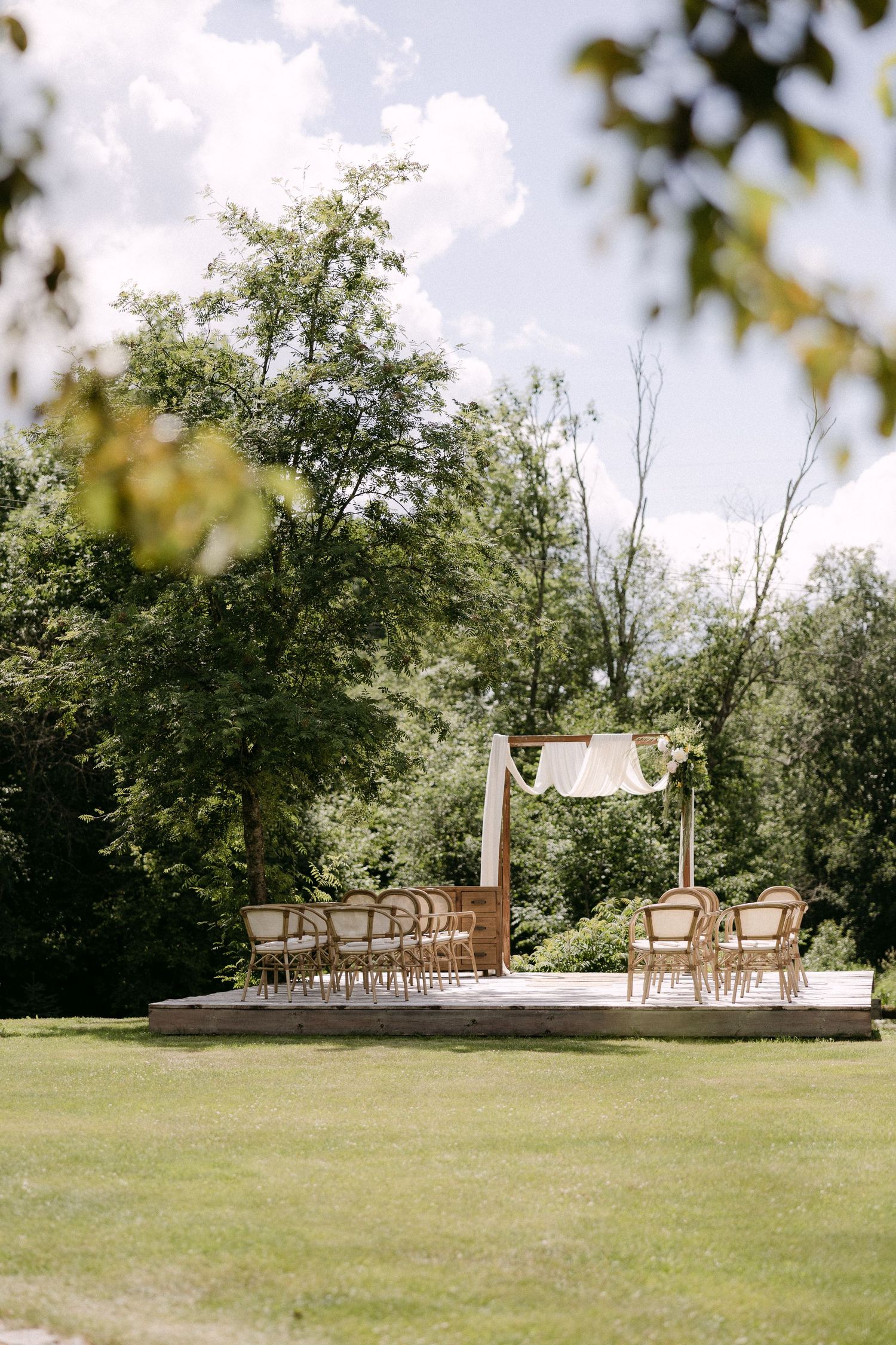 Outdoor wedding ceremony setup with wooden arch, draped fabric, rattan chairs on a platform surrounded by lush green trees.