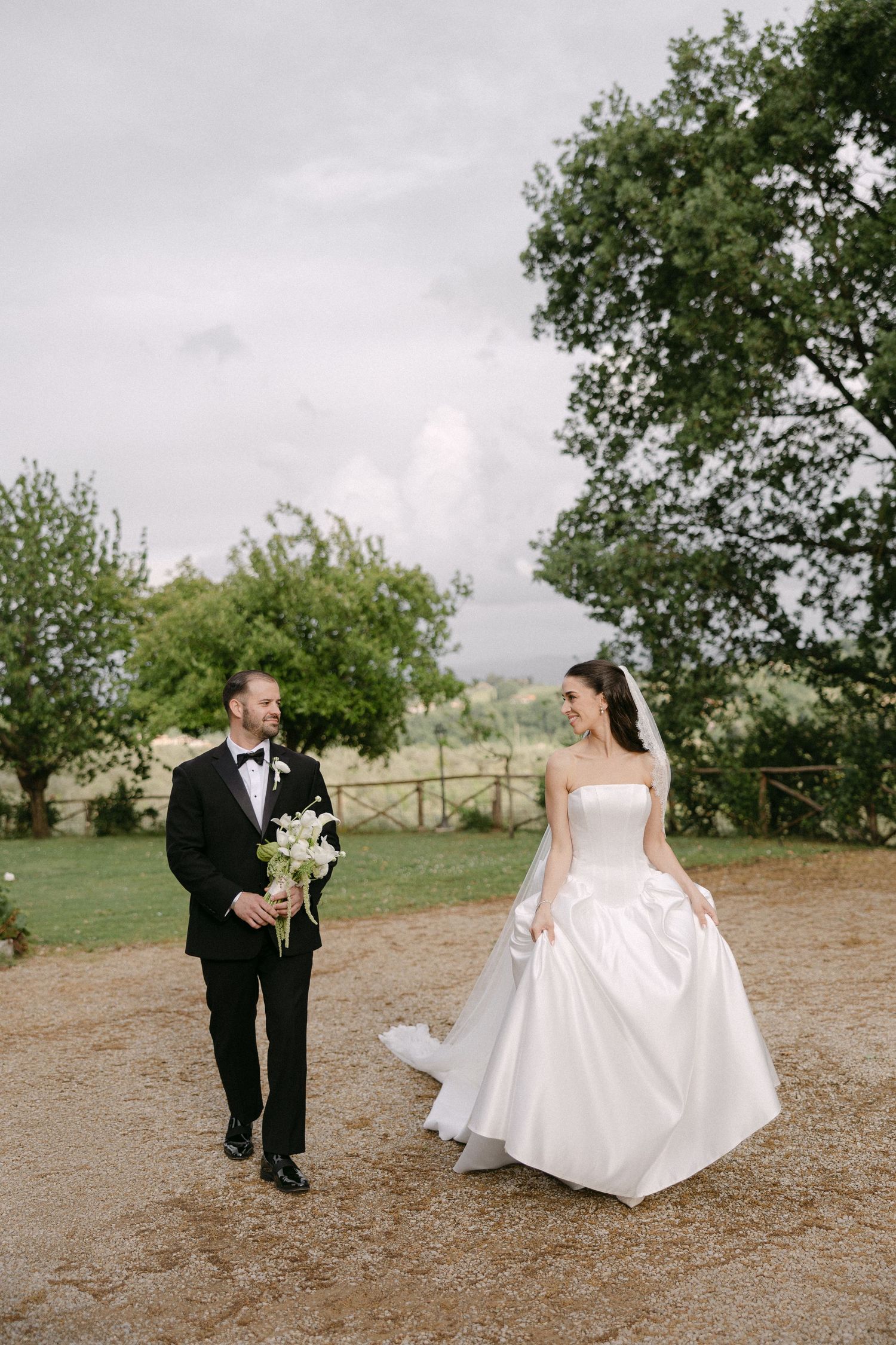 Bride and groom strolling on gravel path outdoors, groom carrying white calla lily bouquet on cloudy day.