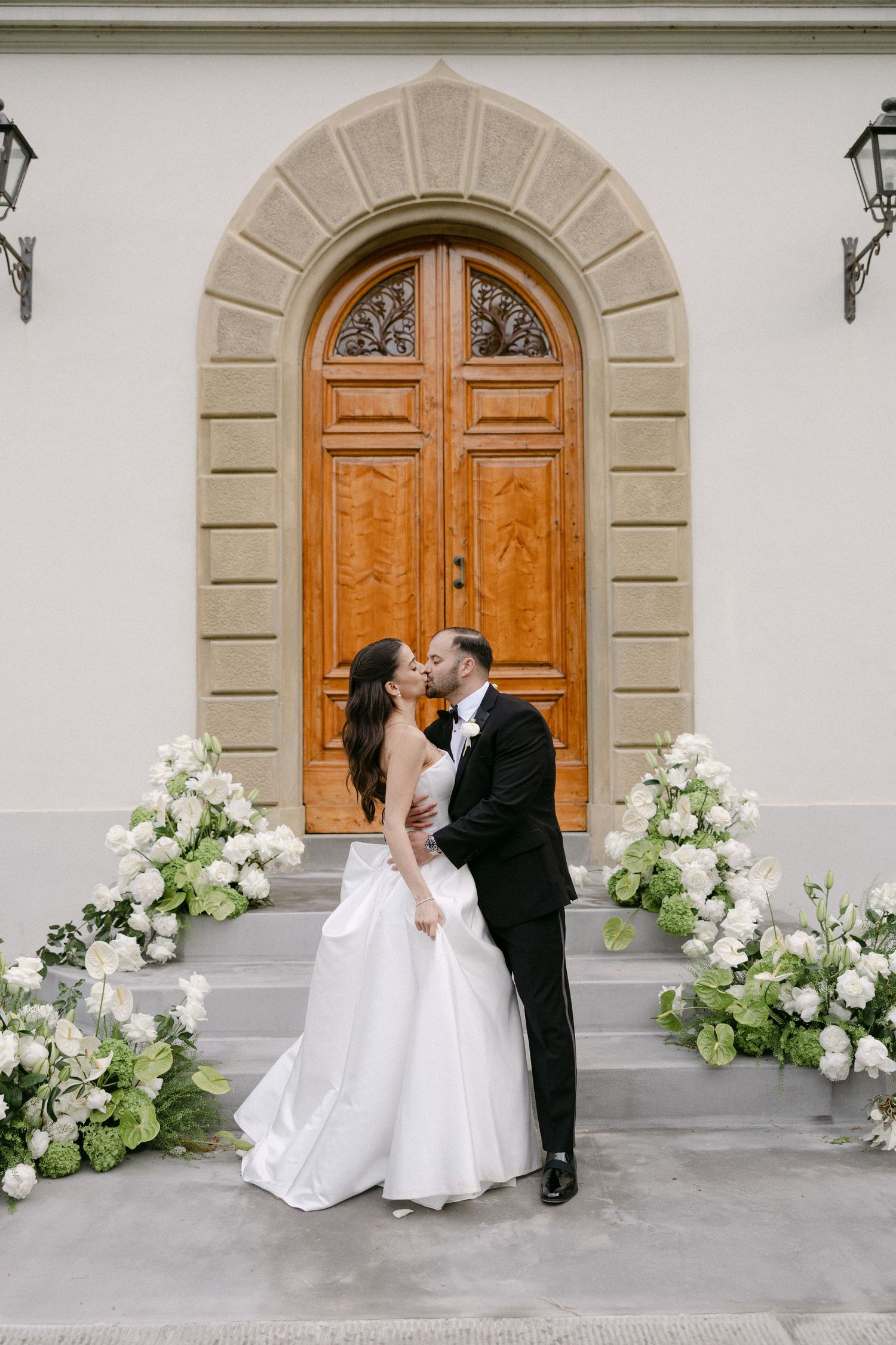 Bride and groom share a kiss on steps flanked by white floral arrangements before elegant arched wooden door.