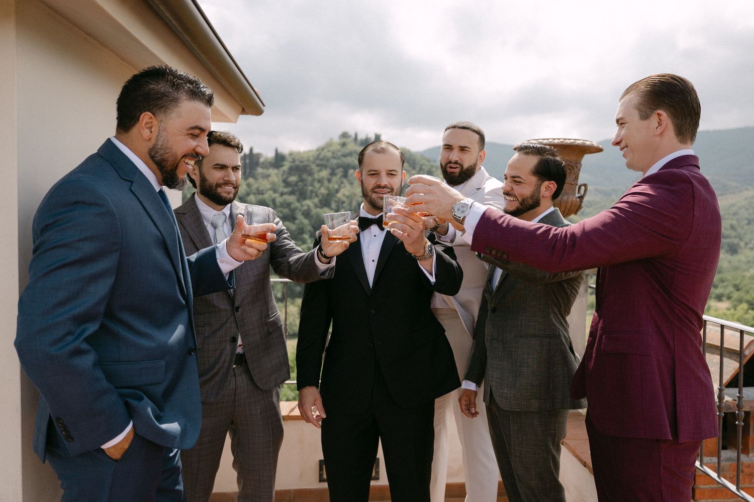Groom and groomsmen in suits toasting with drinks on a balcony with scenic mountain views.