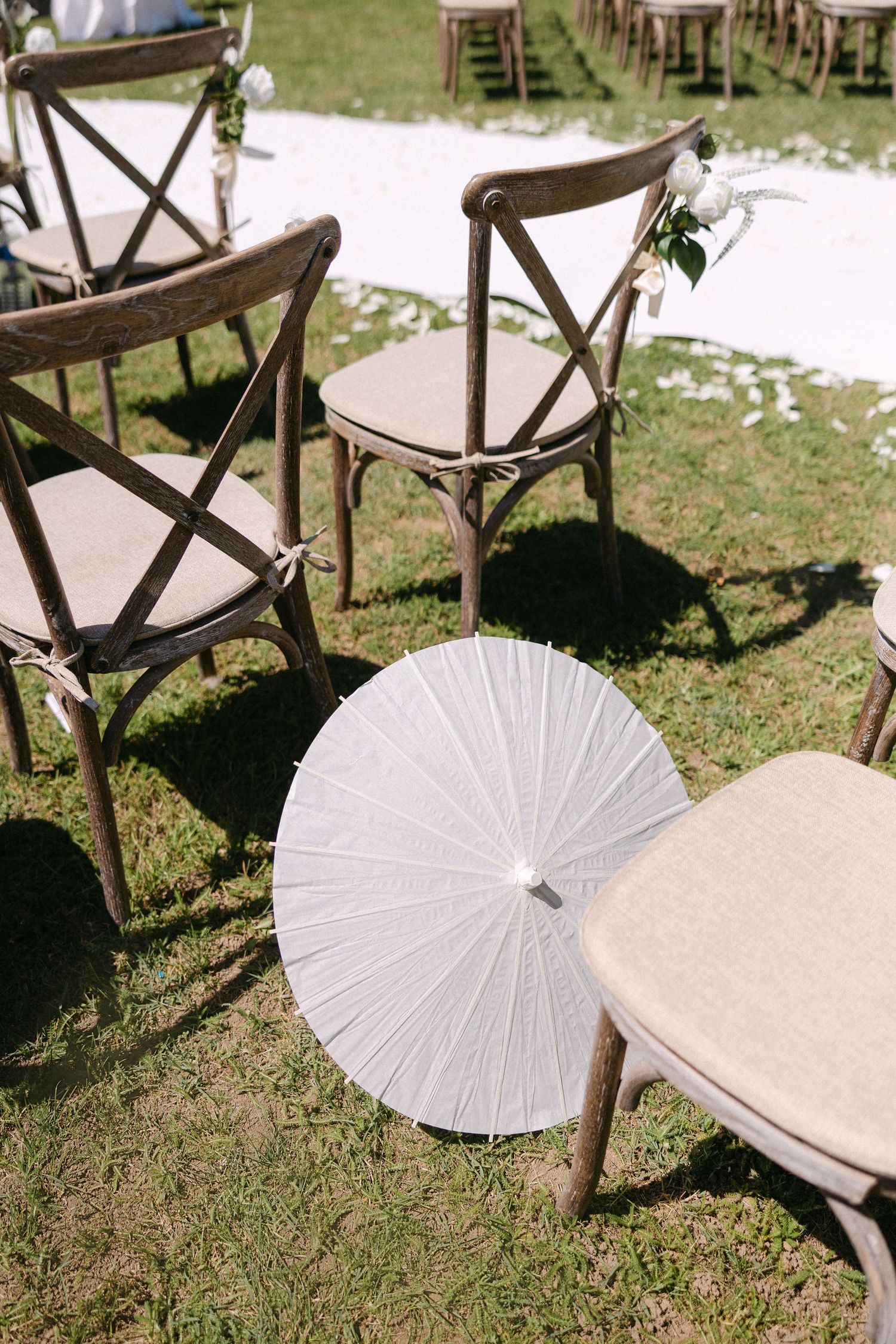 White paper parasol resting against rustic wooden cross-back chairs at outdoor wedding ceremony.