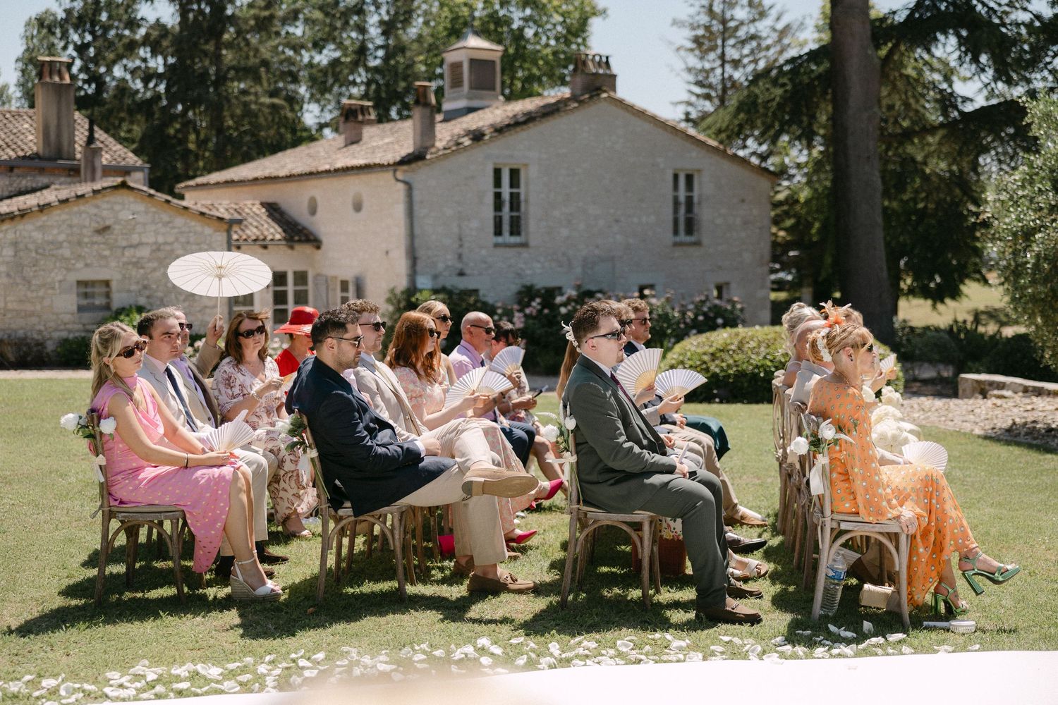 Wedding guests seated outdoors in sunny garden ceremony at a French country estate on a summer day.