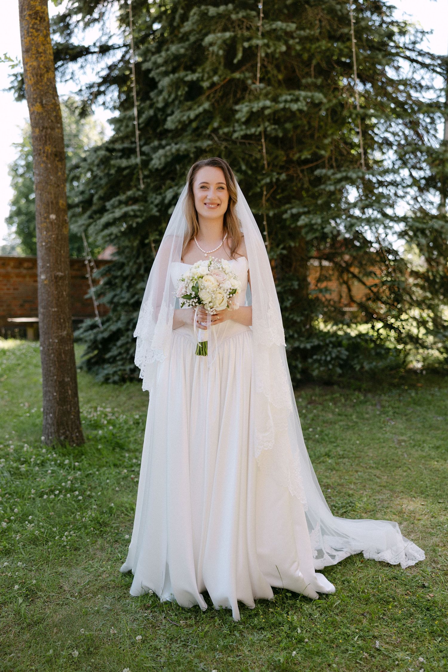 Beautiful bride in white gown and veil holding bouquet in sunny garden setting.