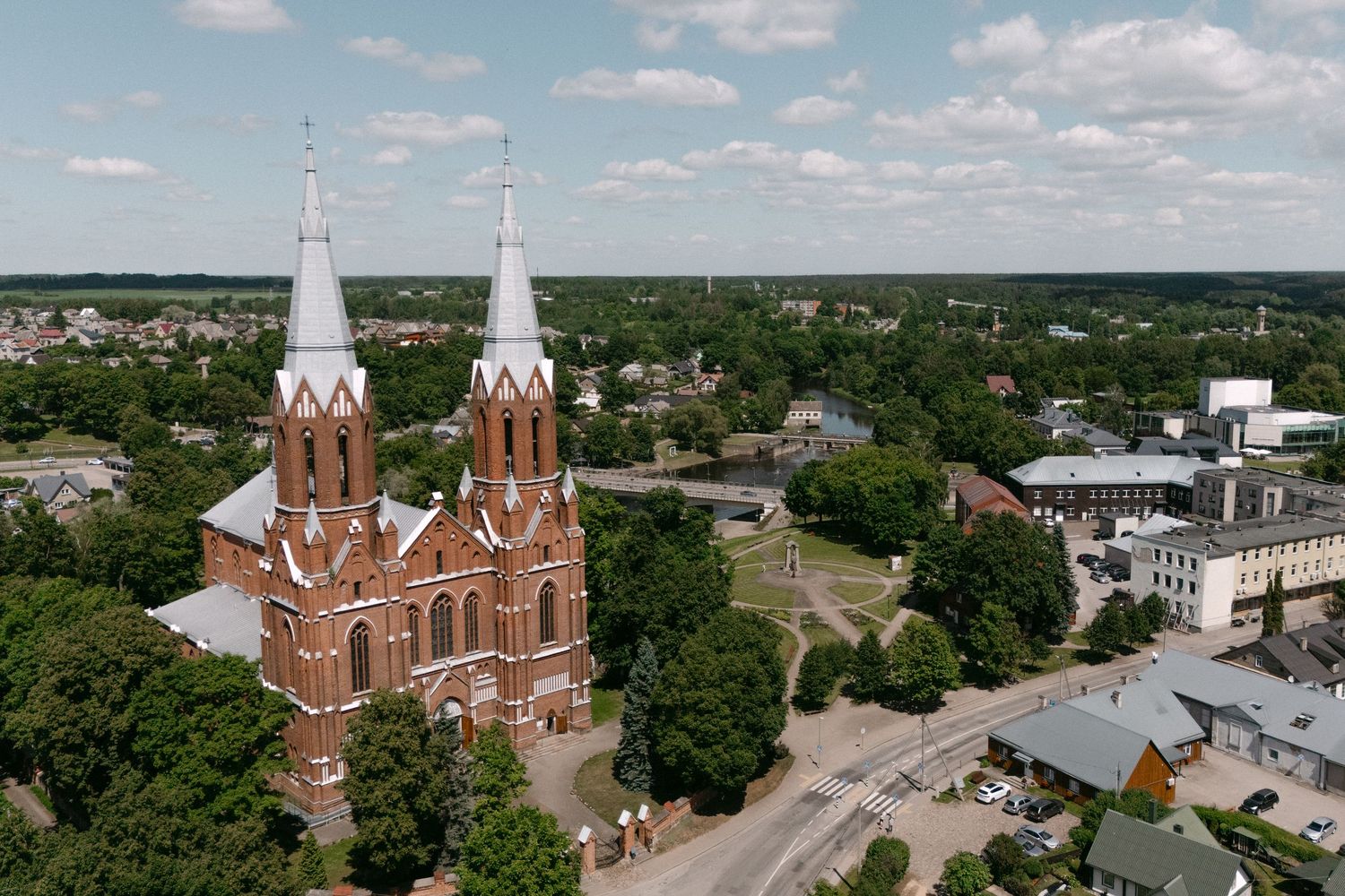 Aerial view of a red brick Gothic church with twin spires surrounded by lush green trees in a small town.