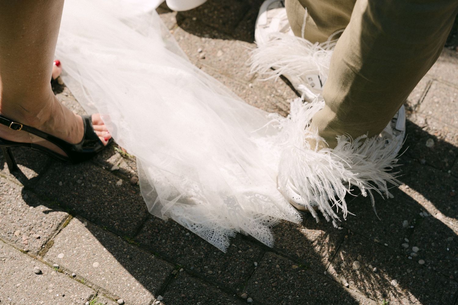 Close-up of a white feathered wedding dress hem on cobblestone, with black heels visible beside it.