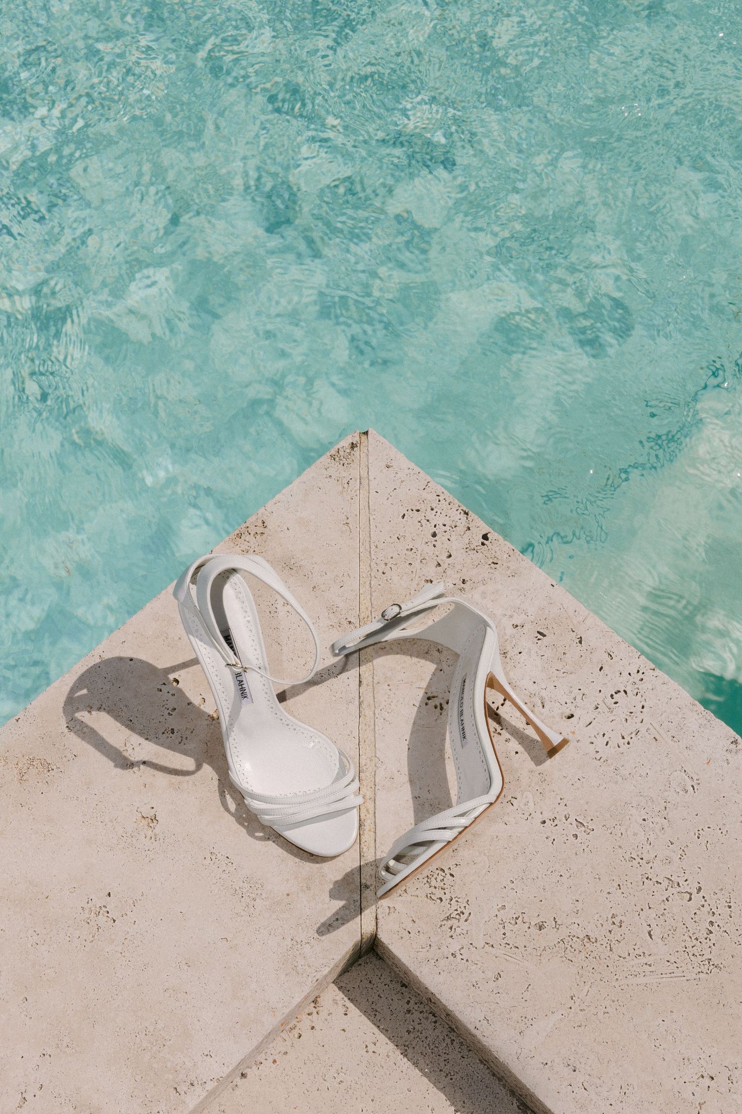 White strappy heeled sandals placed on a marble pool edge with crystal clear turquoise water in the background.