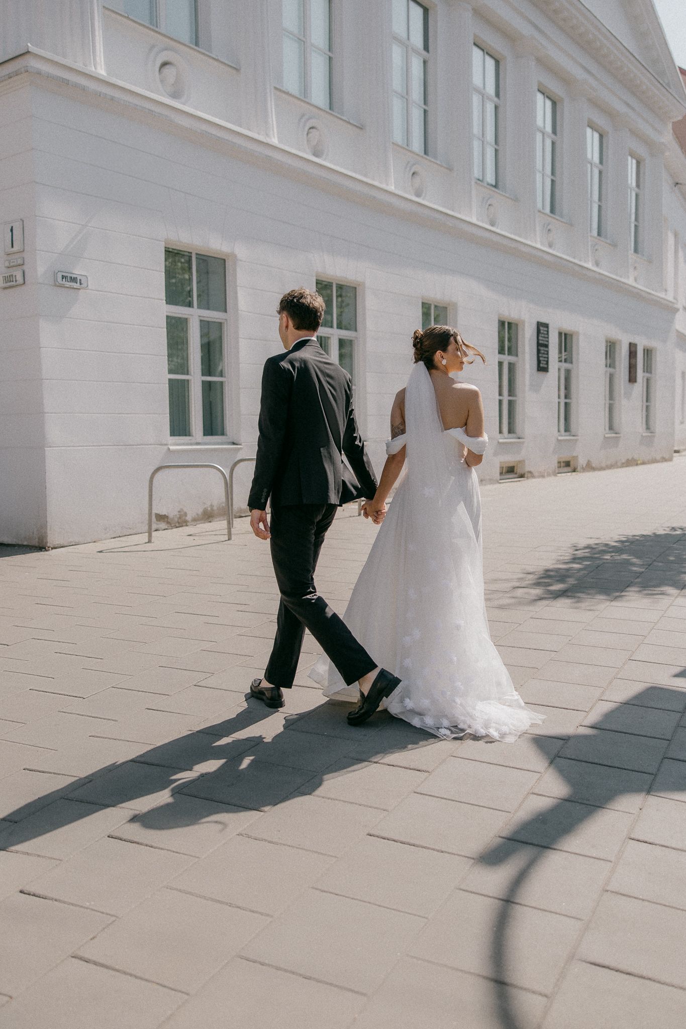 Wedding couple walking along bright white building exterior creating dramatic shadows.