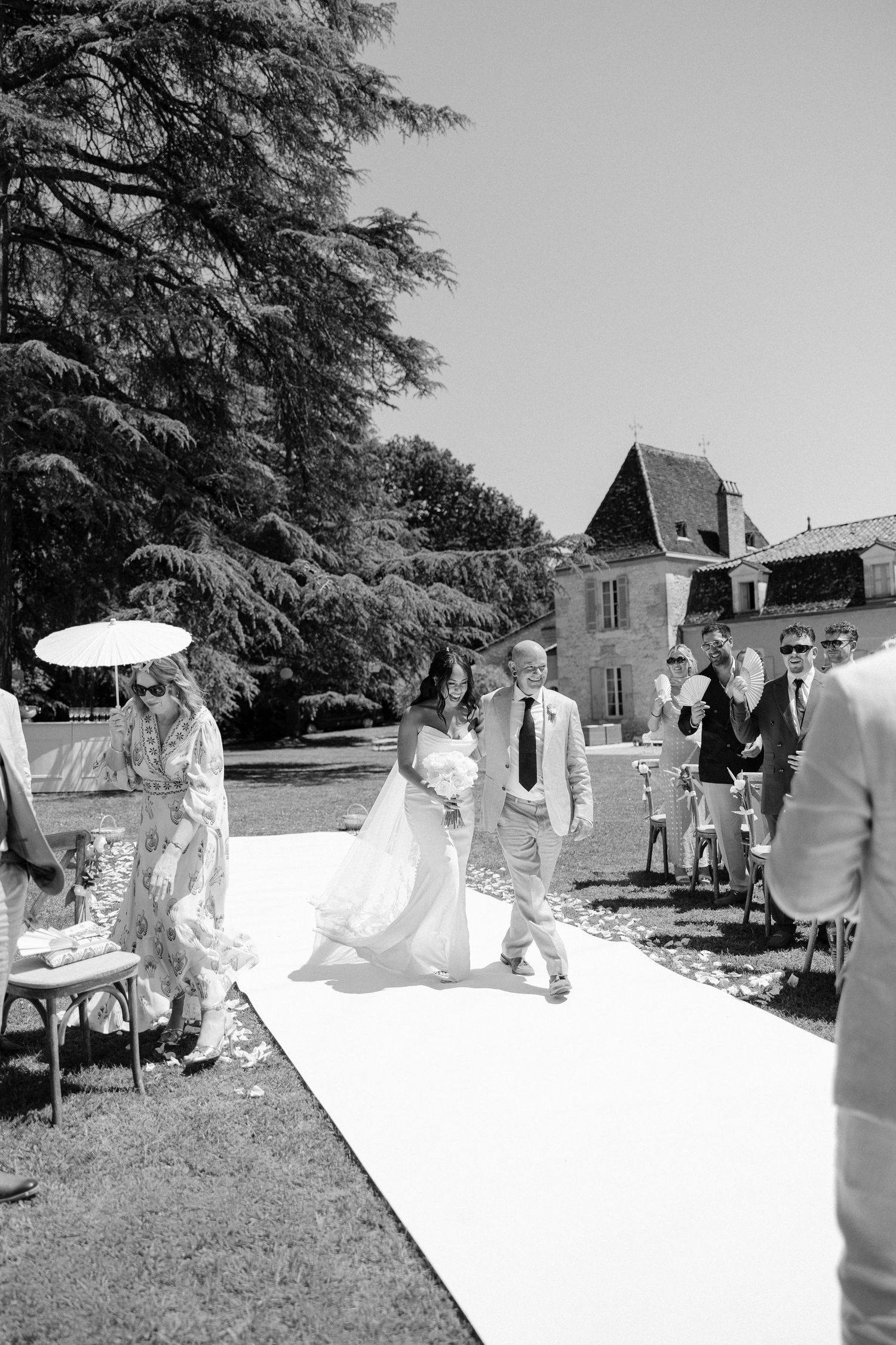 Black and white photo of a bride walking down the aisle at an outdoor wedding ceremony beside a French chateau.