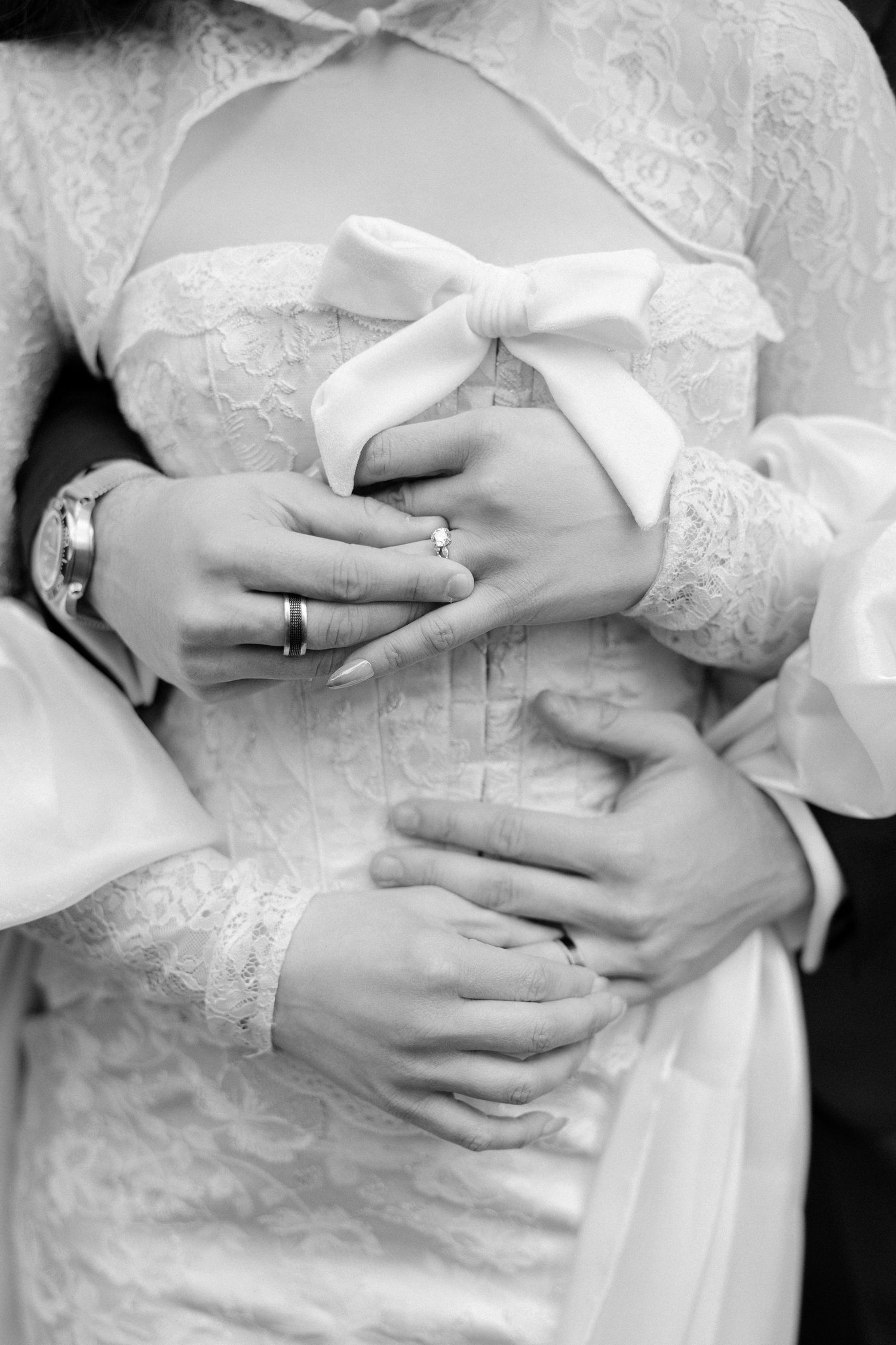 Close-up black and white photo of bride and groom hands embracing, showing wedding rings and lace dress.
