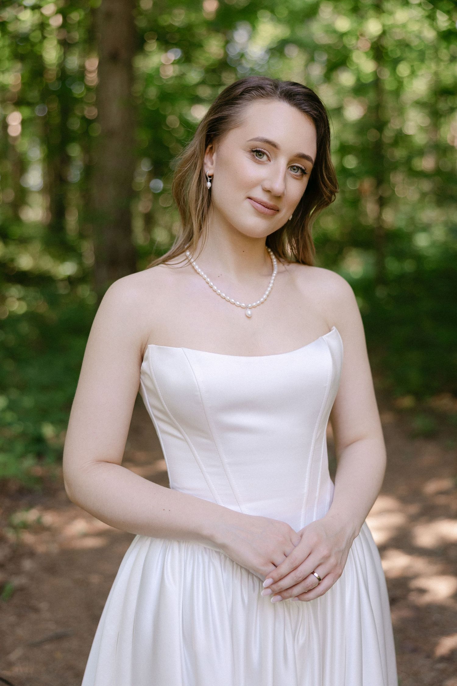 Elegant bride in strapless white gown with pearl necklace posing in sunlit forest setting.