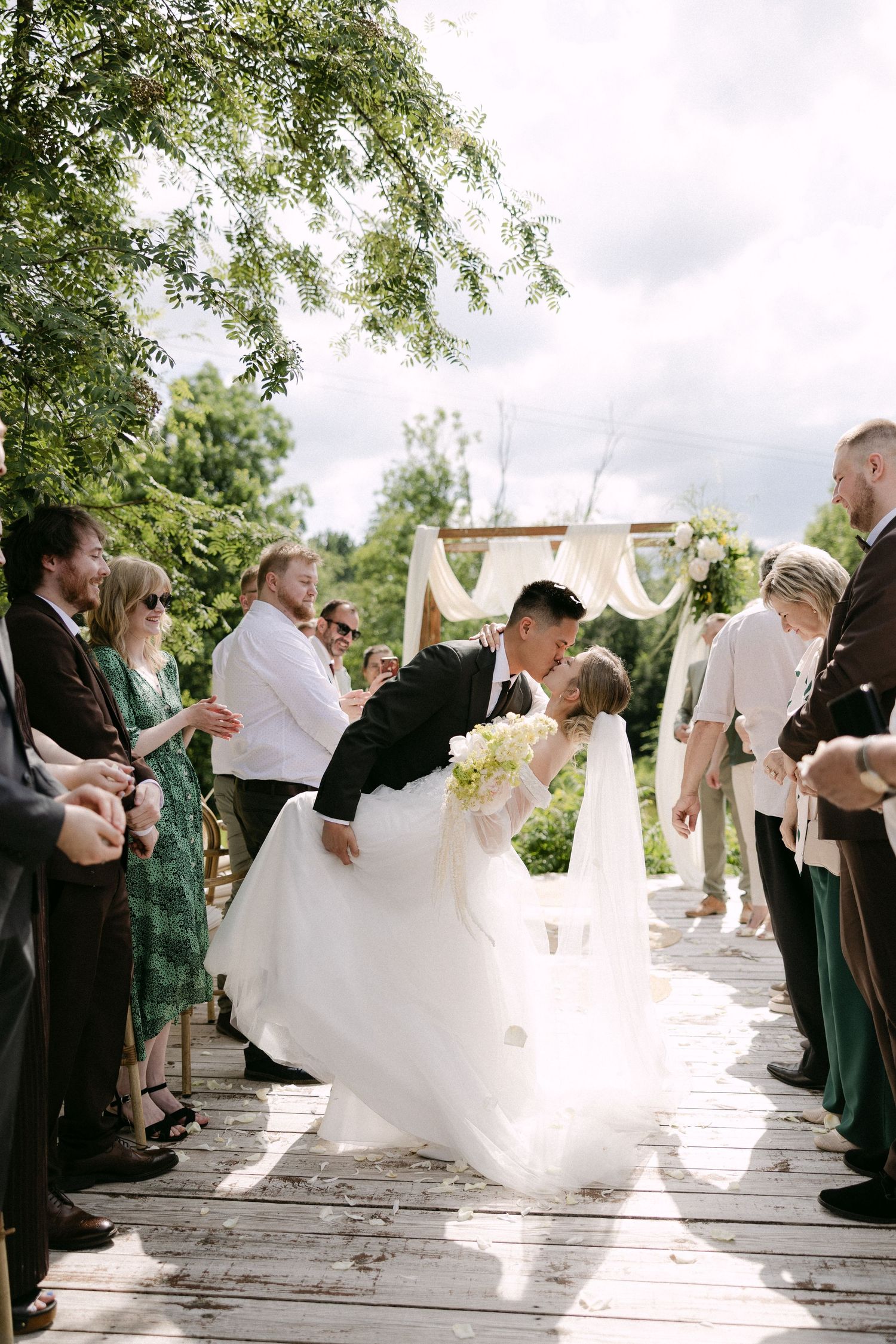 Groom dipping bride for a romantic kiss at outdoor wedding ceremony surrounded by cheering guests.