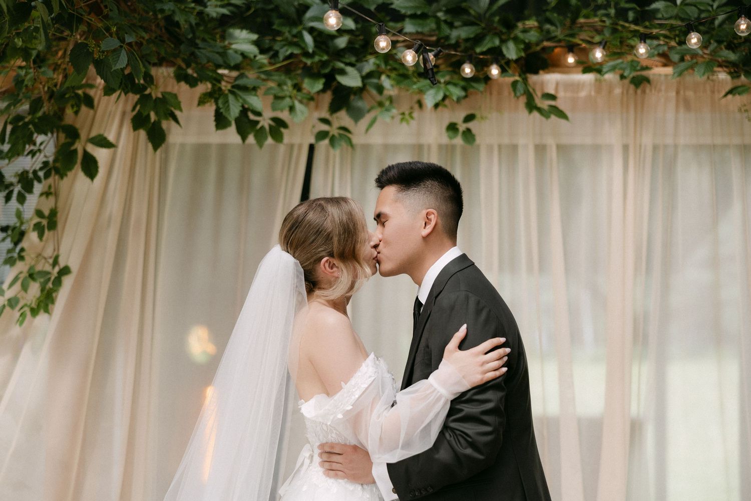 A bride and groom share a romantic first kiss at their elegant wedding ceremony adorned with greenery and string lights.