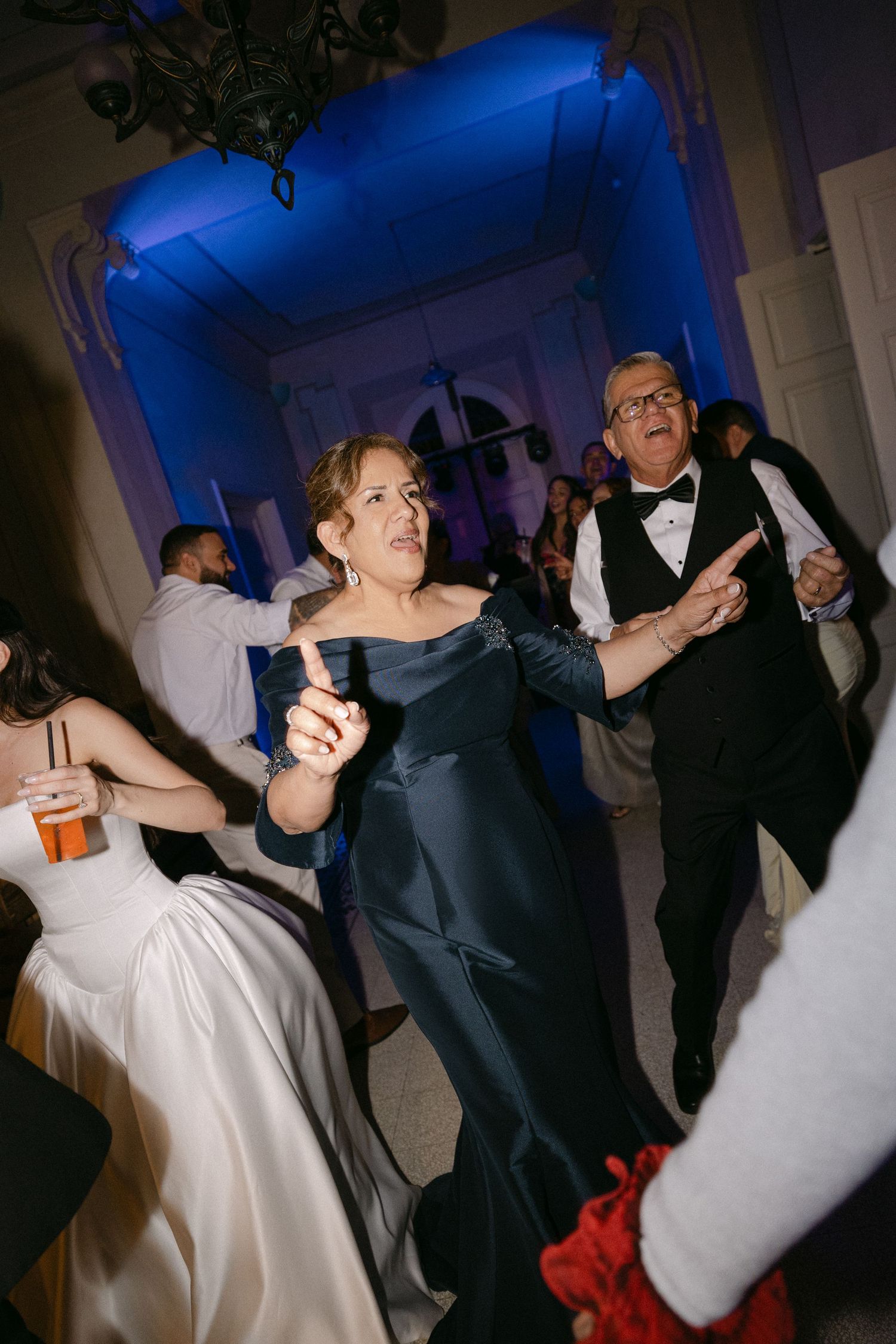 Guests in formal attire dance energetically at wedding reception with blue uplighting ambiance.