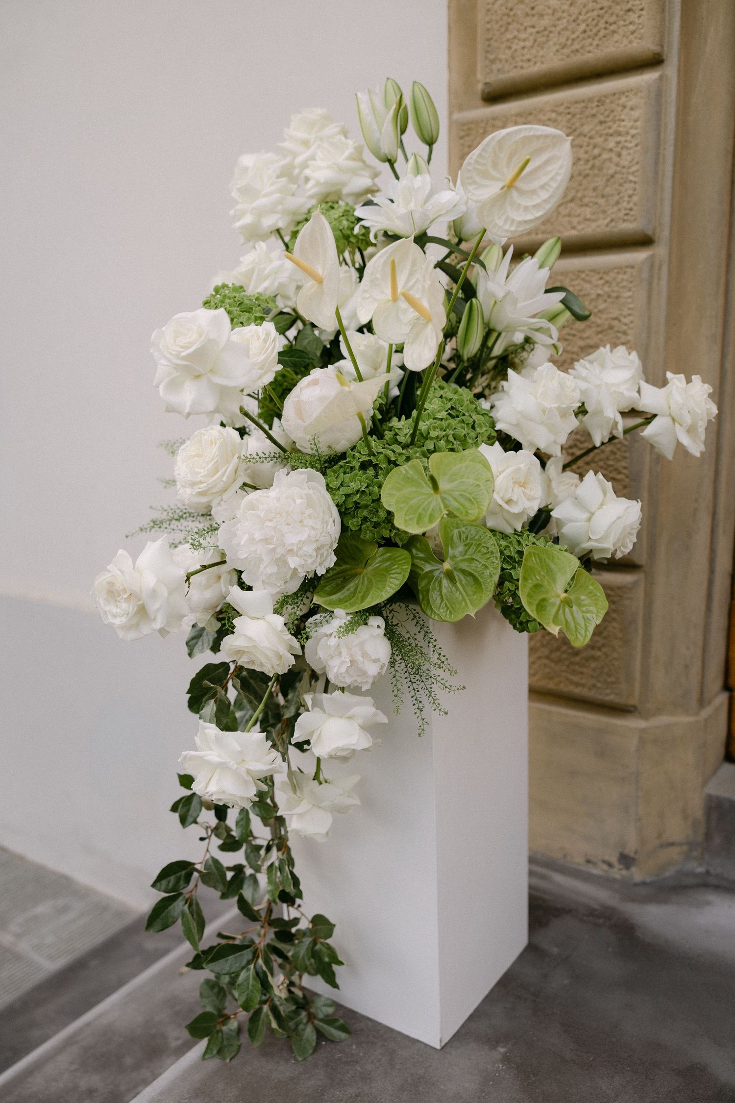 Elegant white wedding floral arrangement with roses, anthuriums, and greenery in a white pedestal vase.