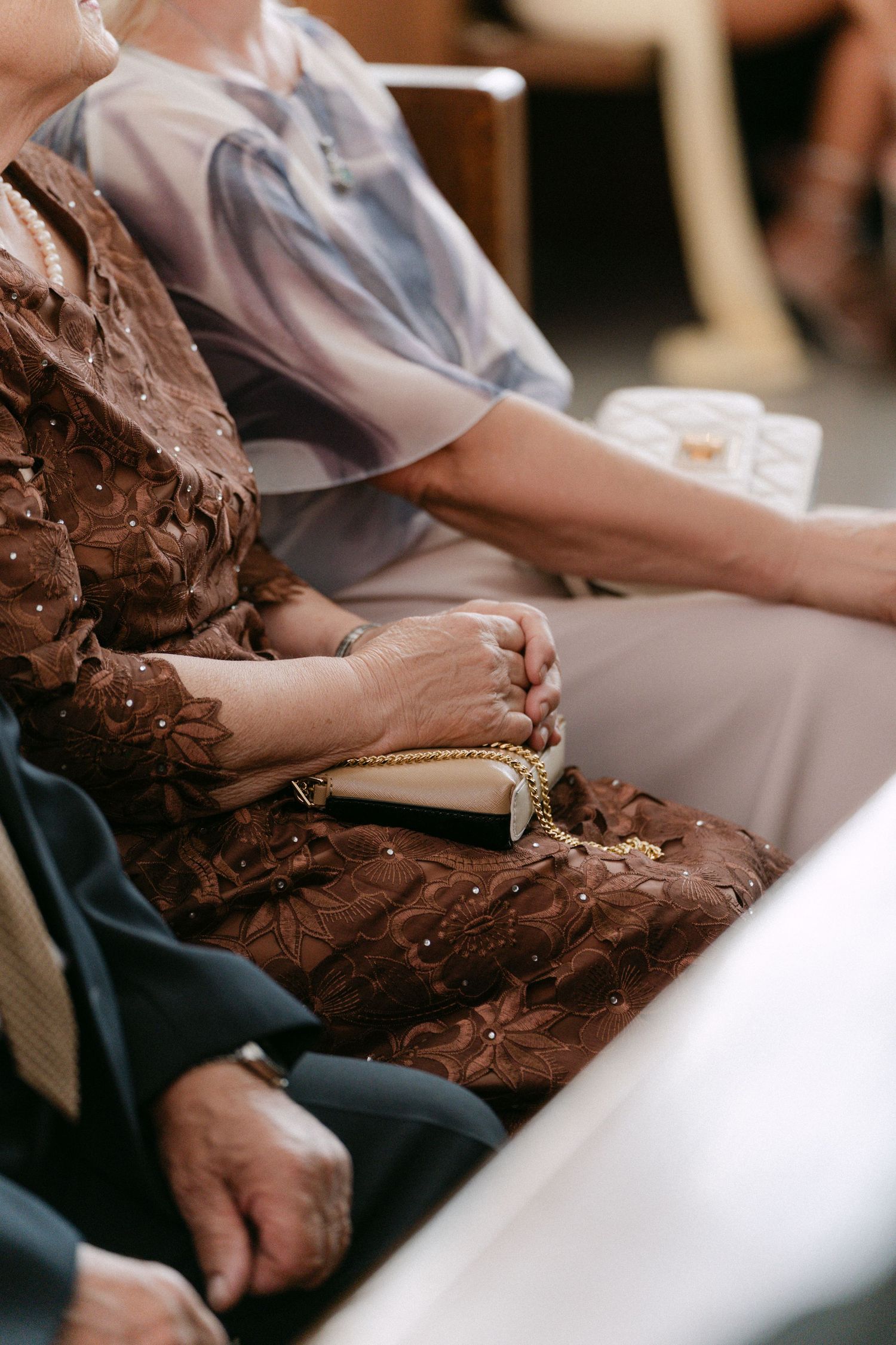 Elderly wedding guests seated in church pews, woman in brown floral dress holding gold clutch purse.
